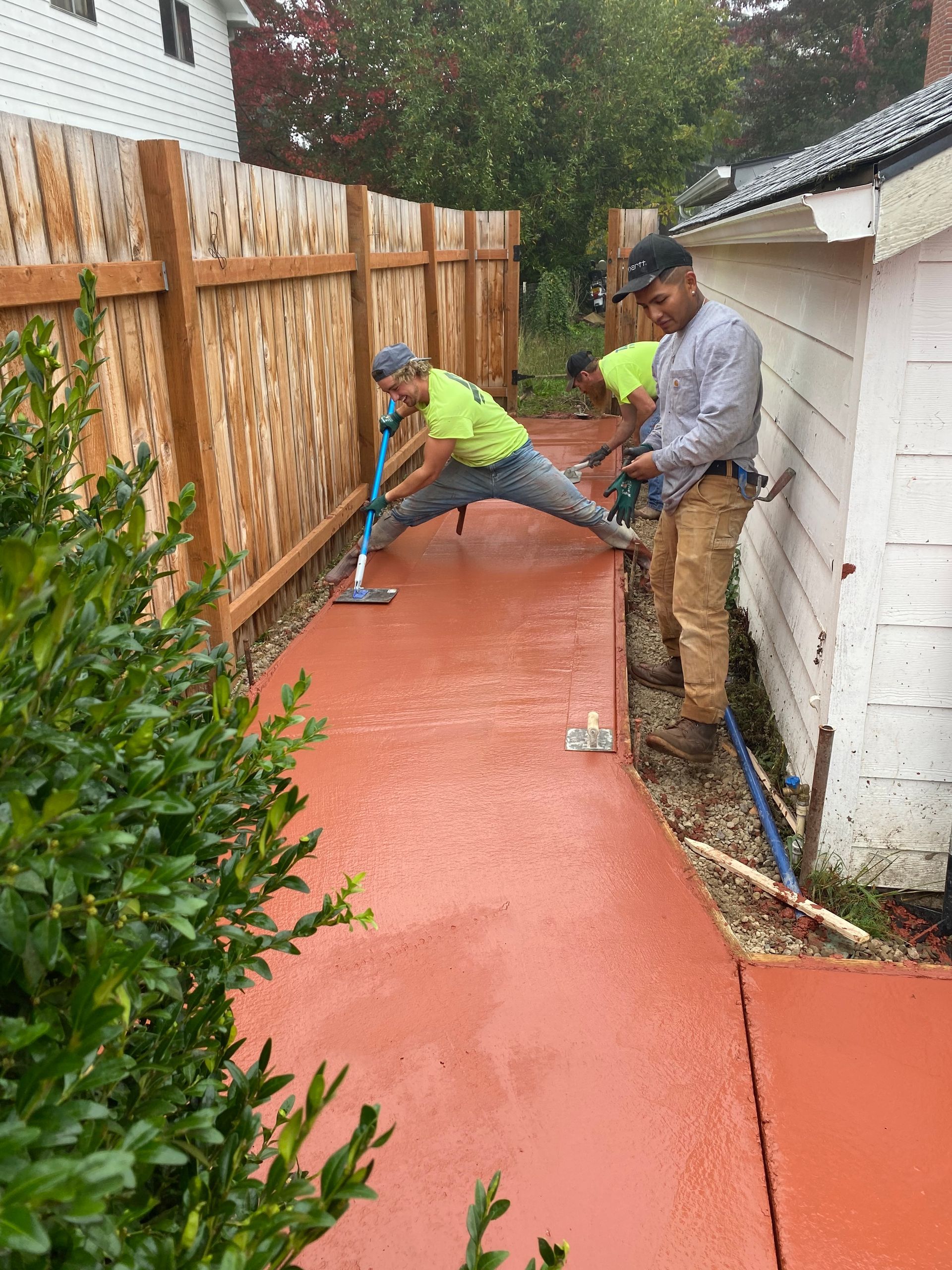 Men smoothing red concrete on a walkway next to a fence and building. One is using a long-handled tool.

