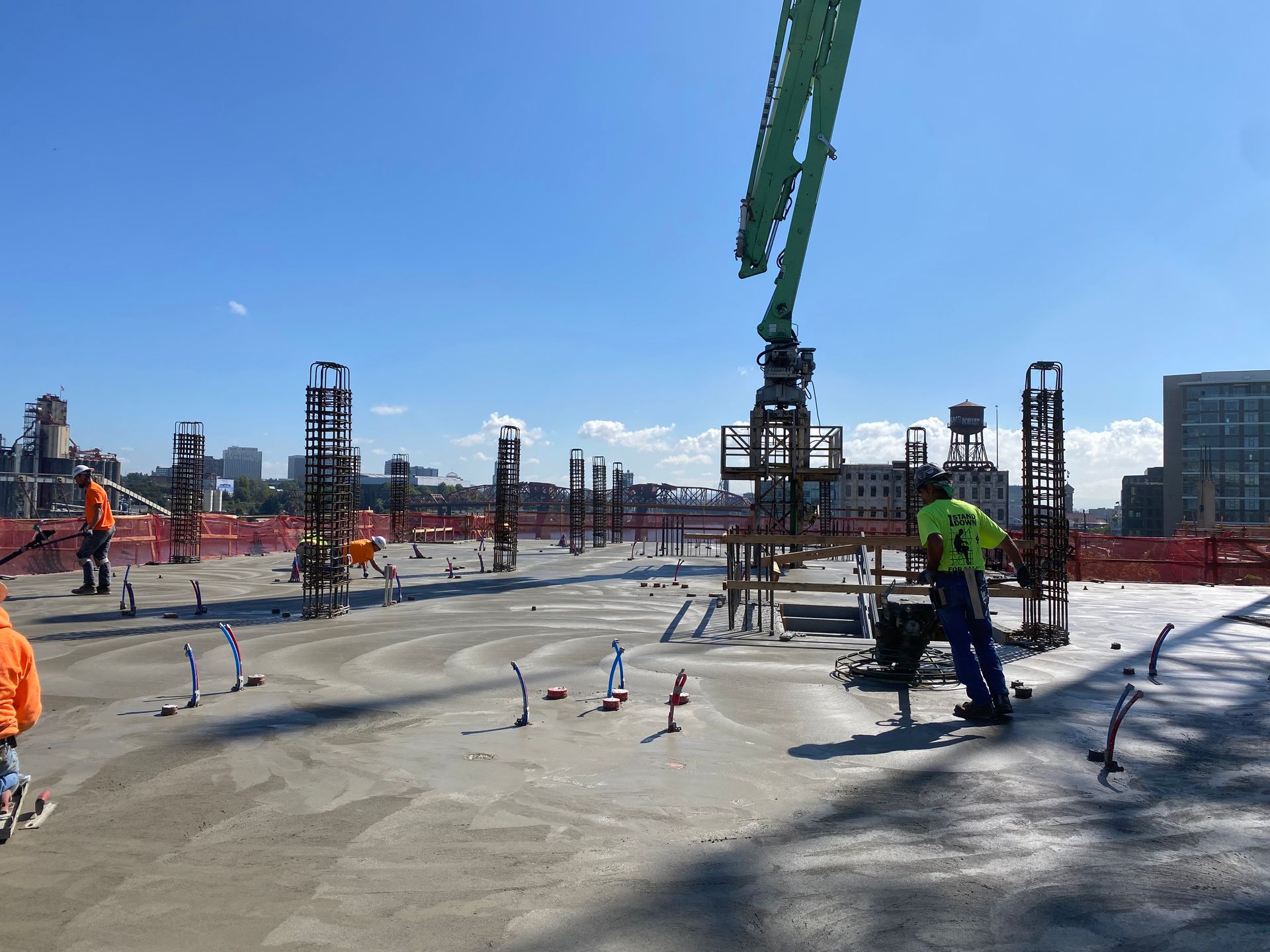 Construction site: concrete being poured onto a deck; workers in safety gear and crane in use; blue sky.