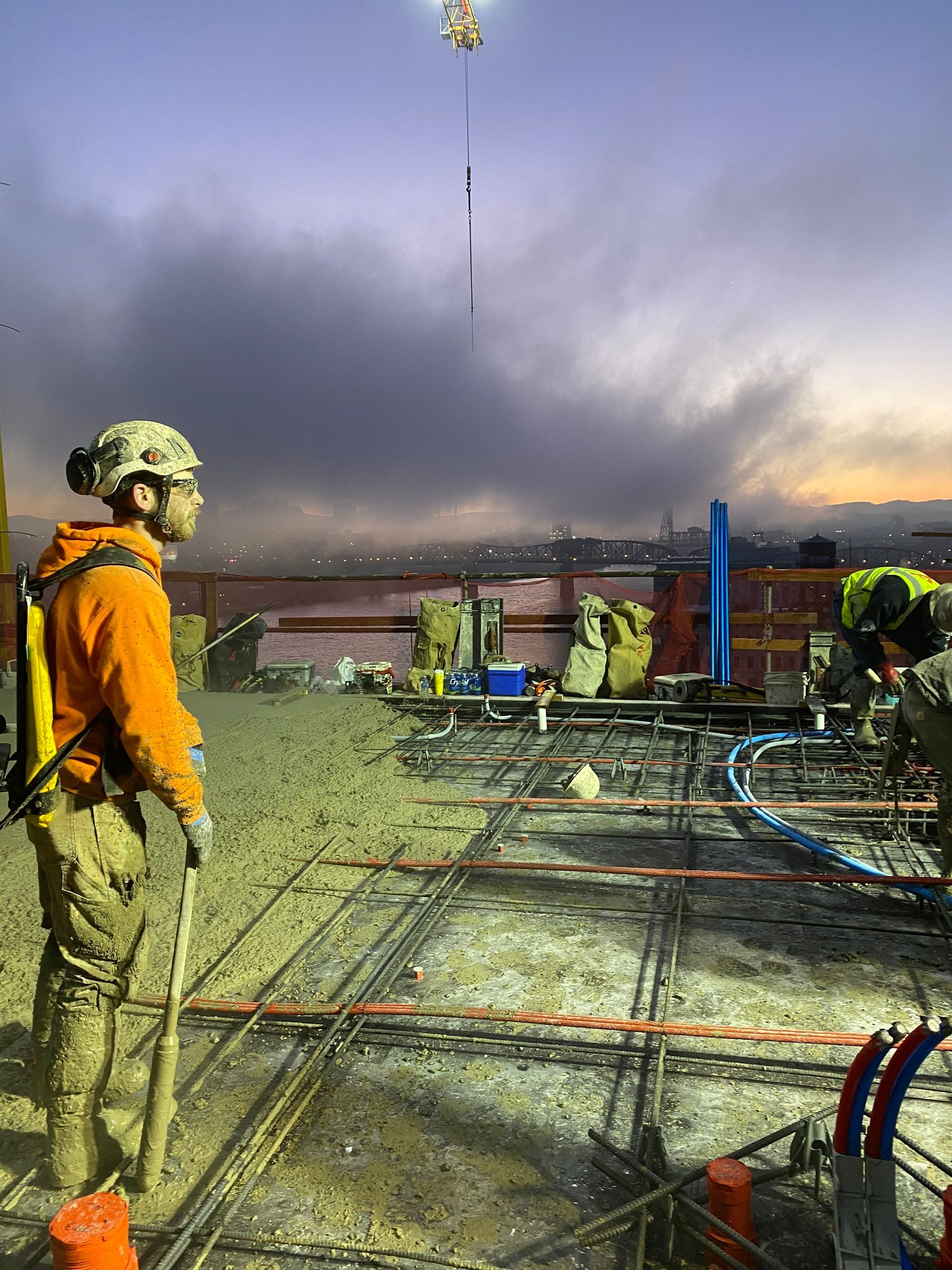 Construction workers on a concrete work site with a basket being lowered from above at dusk.