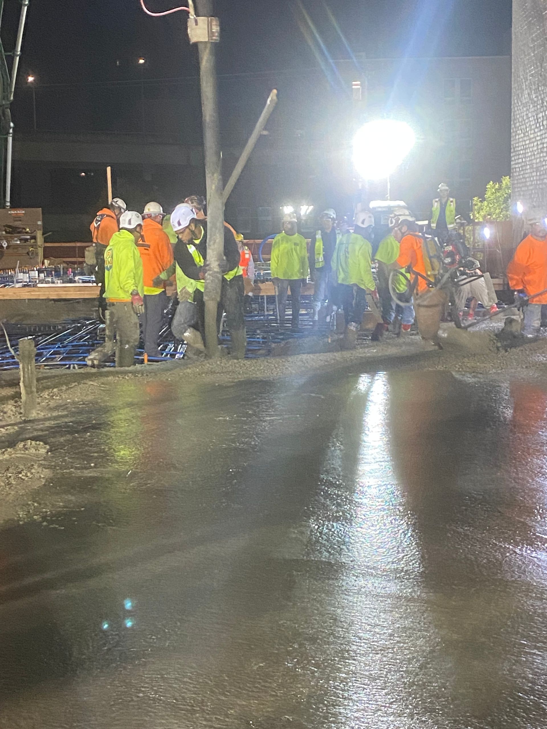 Construction workers in reflective vests and hard hats pour concrete at night under bright lights. Ndamukong Suh