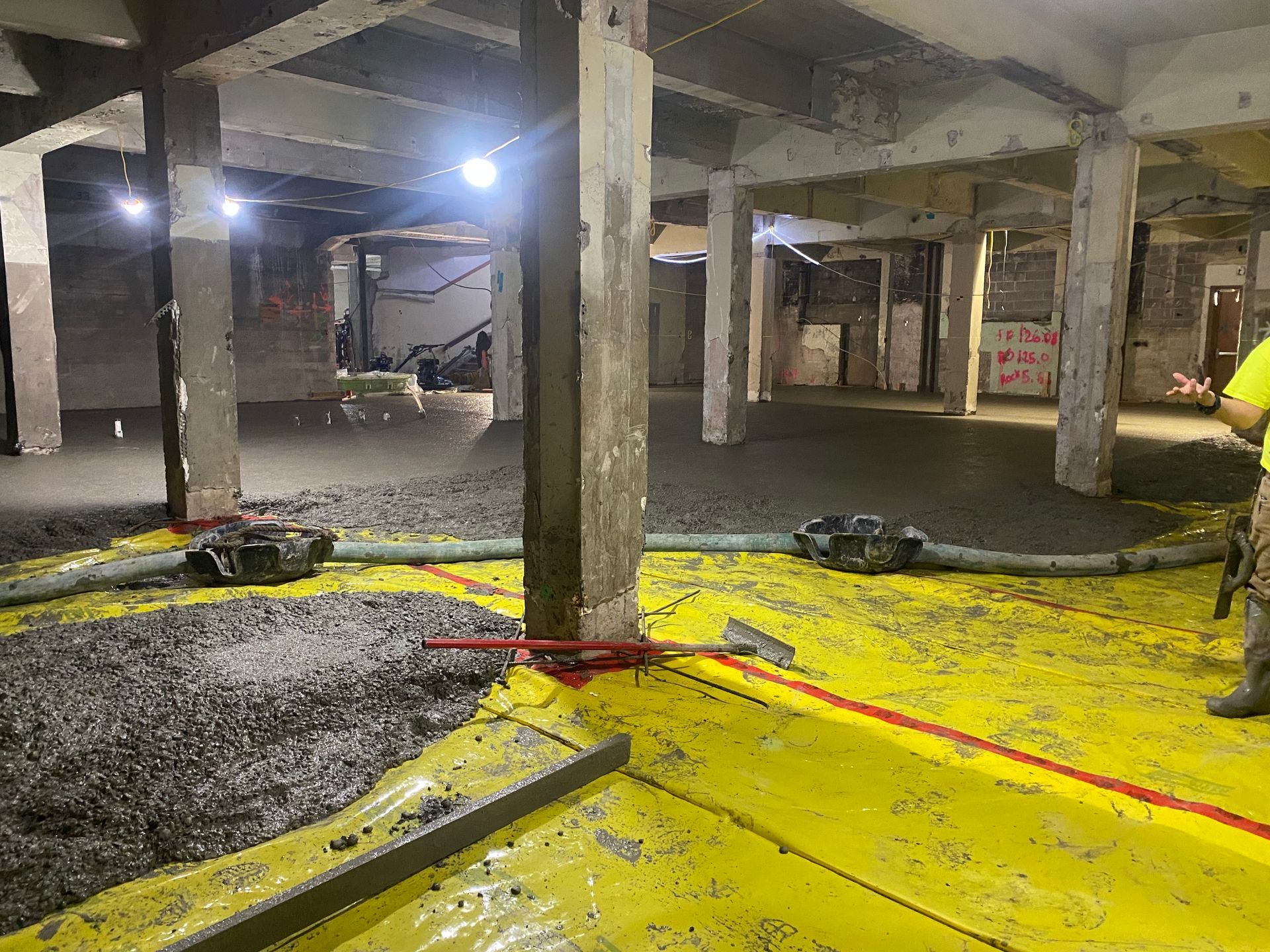 Concrete pouring in a large room with columns; yellow vapor barrier covers the floor, workers visible.