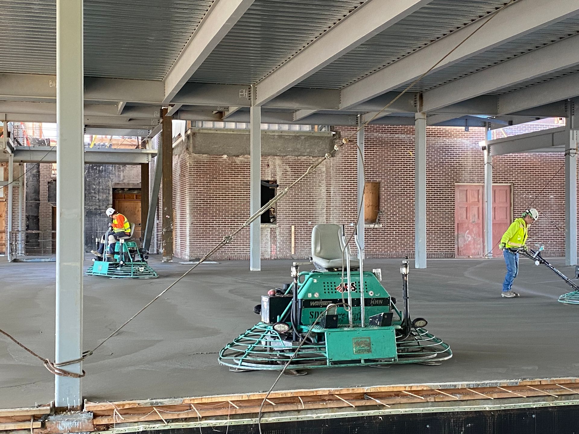 Construction workers using concrete finishing machines on a building floor. Steel beams overhead.