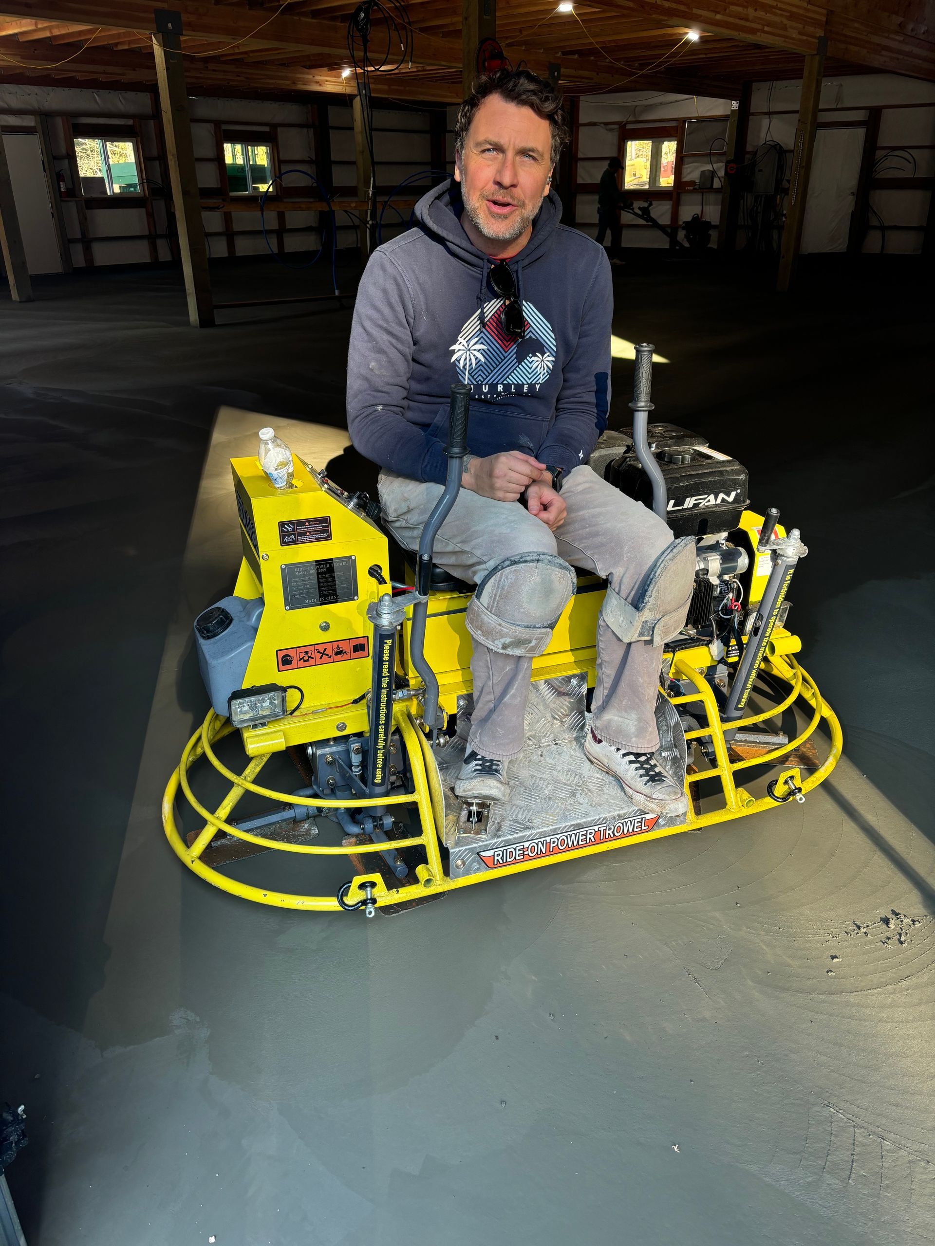 Man operating a yellow concrete finishing machine indoors; he is wearing a gray hoodie and light pants.