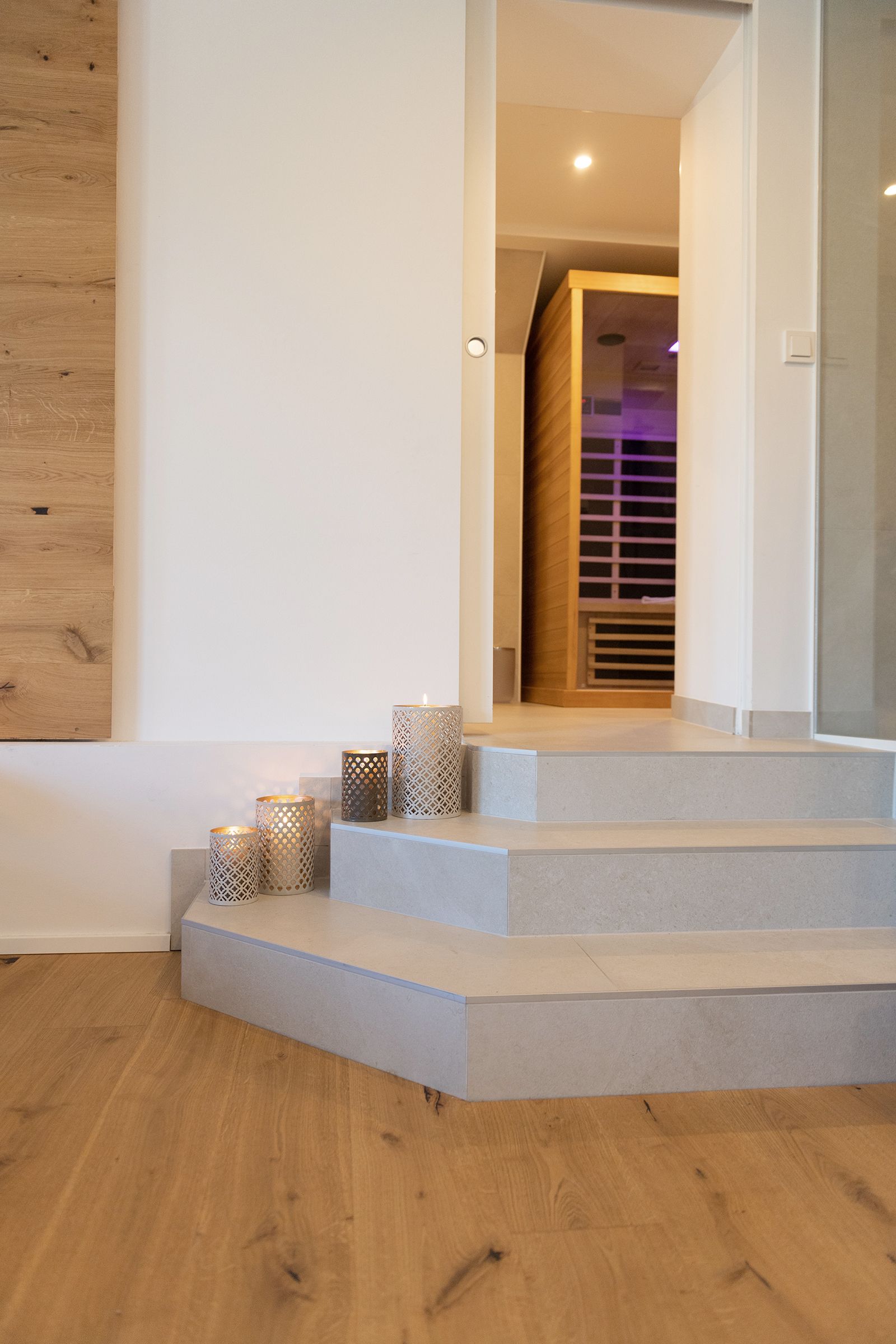 Graue Stufen geschmückt mit Kerzen führen in das Badezimmer und zu der Infrarot Kabine, Brauner Parkettboden und die Hälfte der hölzernen Infrarotkabine ist zu sehen.