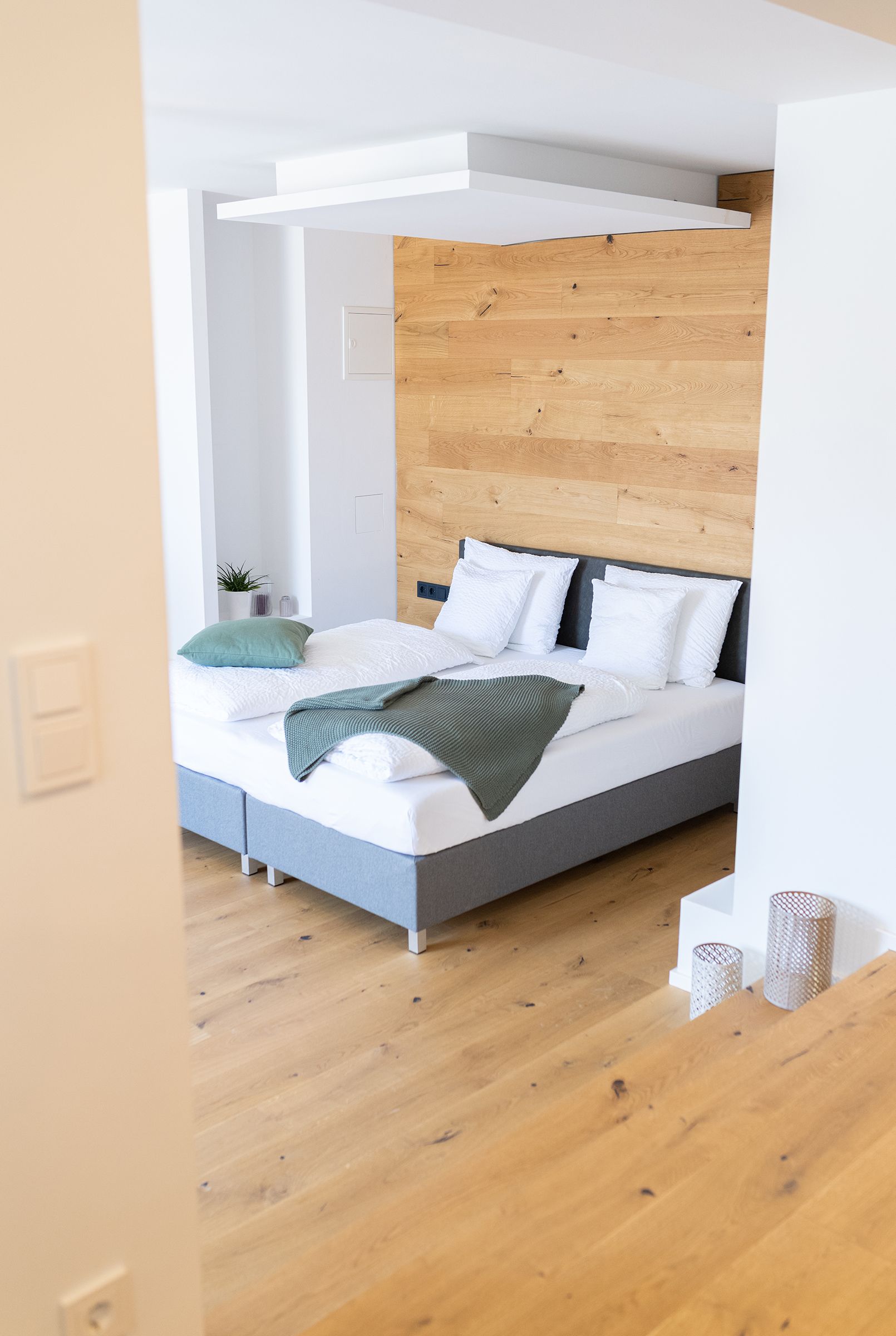 Das graue doppel-Boxspringbett mit weißen Laken steht im Fokus. Der Boden ist Parkett. Das Bett steht mit Kopfteil zu einer holzverkleideten Wand.
