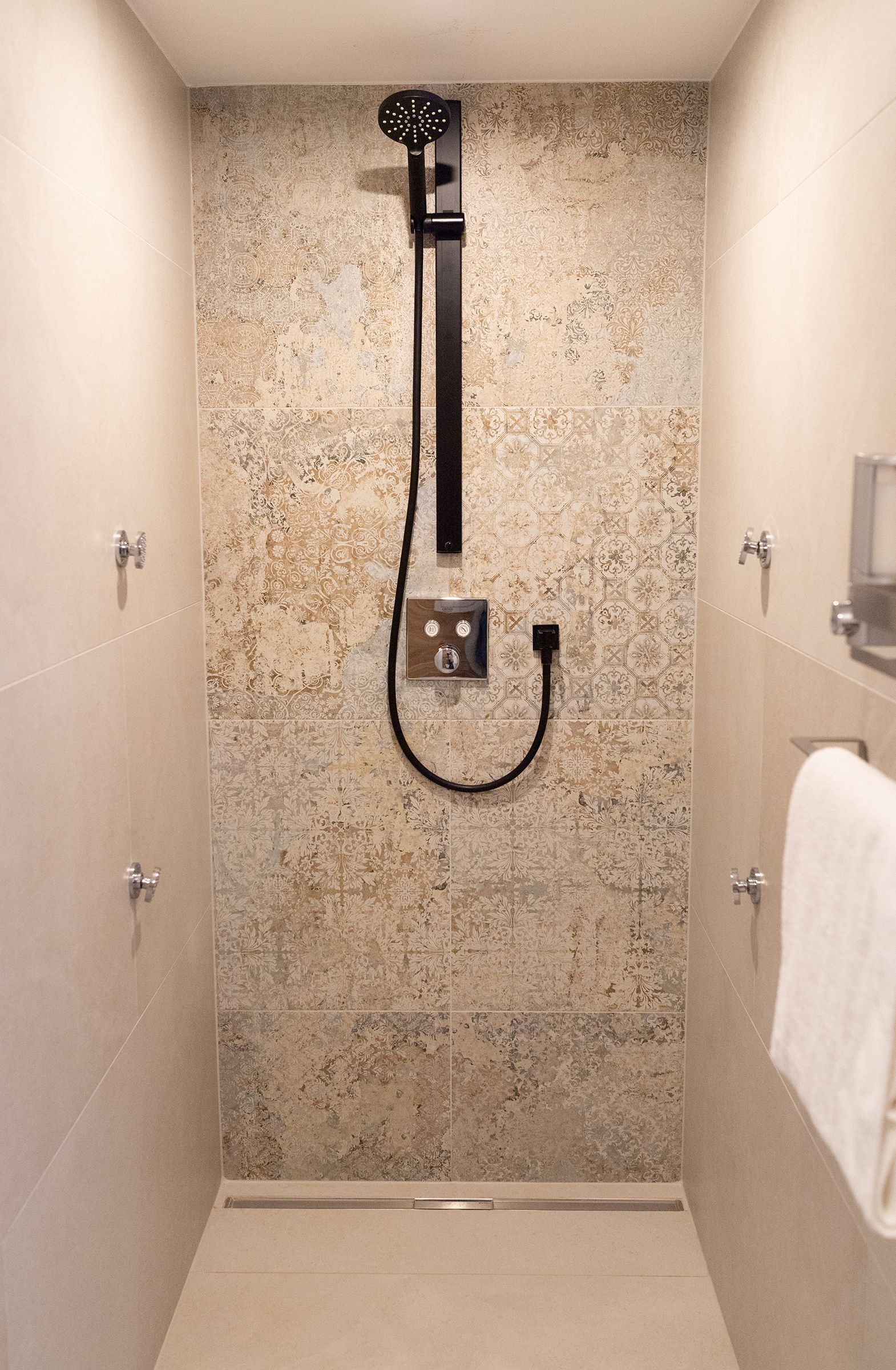Moderne begehbare Dusche mit großer Handbrause und schwarzer Armatur. Die Rückwand ist mit strukturierten, beige-grauen Fliesen im Vintage-Dekor gestaltet, die Seitenwände und der Boden in hellen Naturtönen gehalten.