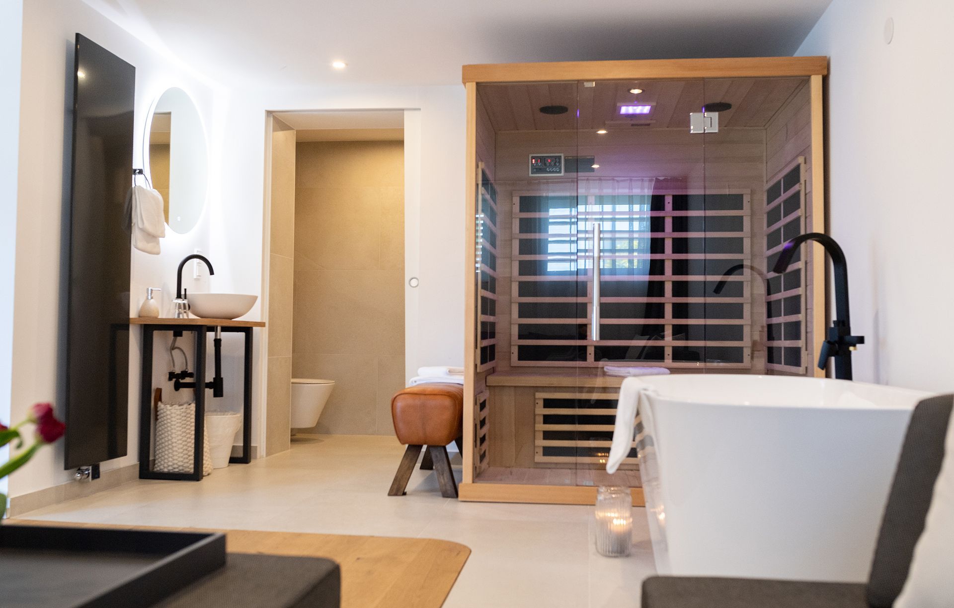 Offenes Badezimmer mit Durchgang zur  vom Raum abgetrennter Toilette. rechts neben der Türe befindet sich die Infrarotkabine. Davor ist die freistehende Badewanne. links neben der Türe ist ein rundes Waschbecken darüber ein großer runder Spiegel.
