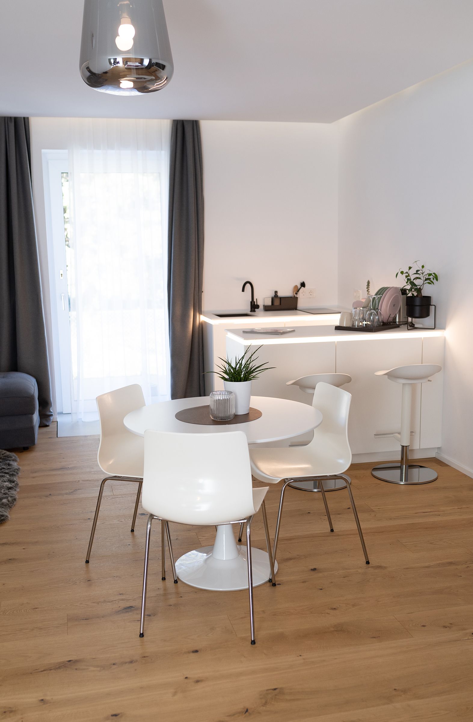 im Raum steht ein weißer runder Tisch mit drei passenden Stühlen, dahinter eine U-förmige weiße Küchenzeile. Gegenüber der Küche ist eine graue L-förmige Couch und ein kleiner Teppich in der selben Farbe. Die große Fenstertüre ist offen