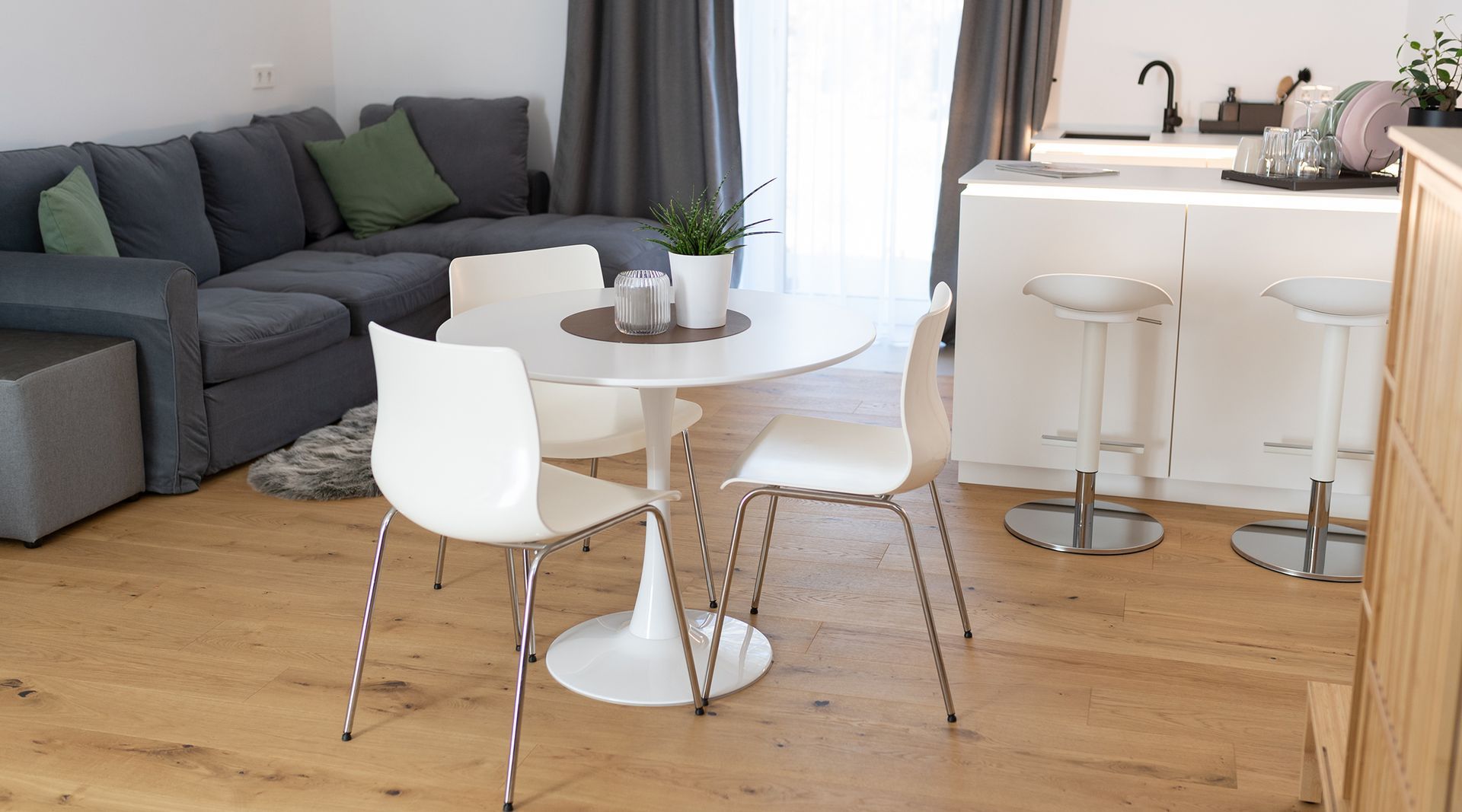 im Raum steht ein weißer runder Tisch mit drei passenden Stühlen, dahinter eine U-förmige weiße Küchenzeile. Gegenüber der Küche ist eine graue L-förmige Couch und ein kleiner Teppich in der selben Farbe. Die große Fenstertür und grauen Vorhänge sind offen.