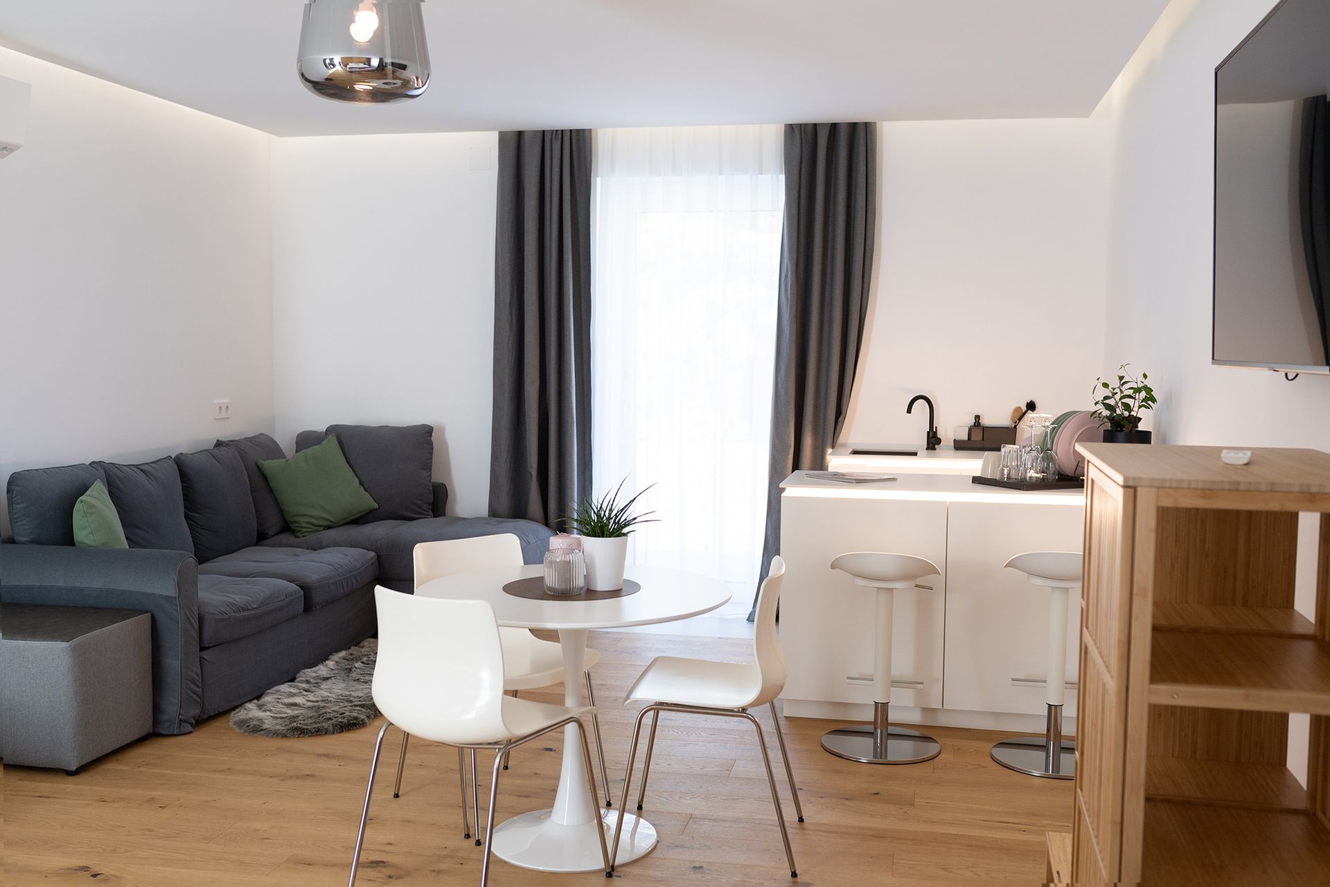 im Raum steht ein weißer runder Tisch mit drei passenden Stühlen, dahinter eine U-förmige weiße Küchenzeile. Gegenüber der Küche ist eine graue L-förmige Couch und ein kleiner Teppich in der selben Farbe. Die große Fenstertüre und grauen Vorhänge sind offen.