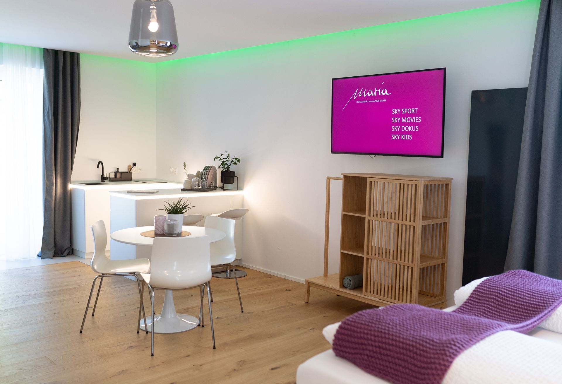 Holzparkettboden und weiße Wände mit grüner indirekten Beleuchtung an dem Rand der Decke. Ein Fernseher hängt an der Wand. darunter ist eine moderne Kommode aus Holz. im Raum steht ein weißer runder Tisch mit drei Passenden Stühlen, dahinter eine U-förmige weiße Küchenzeile.