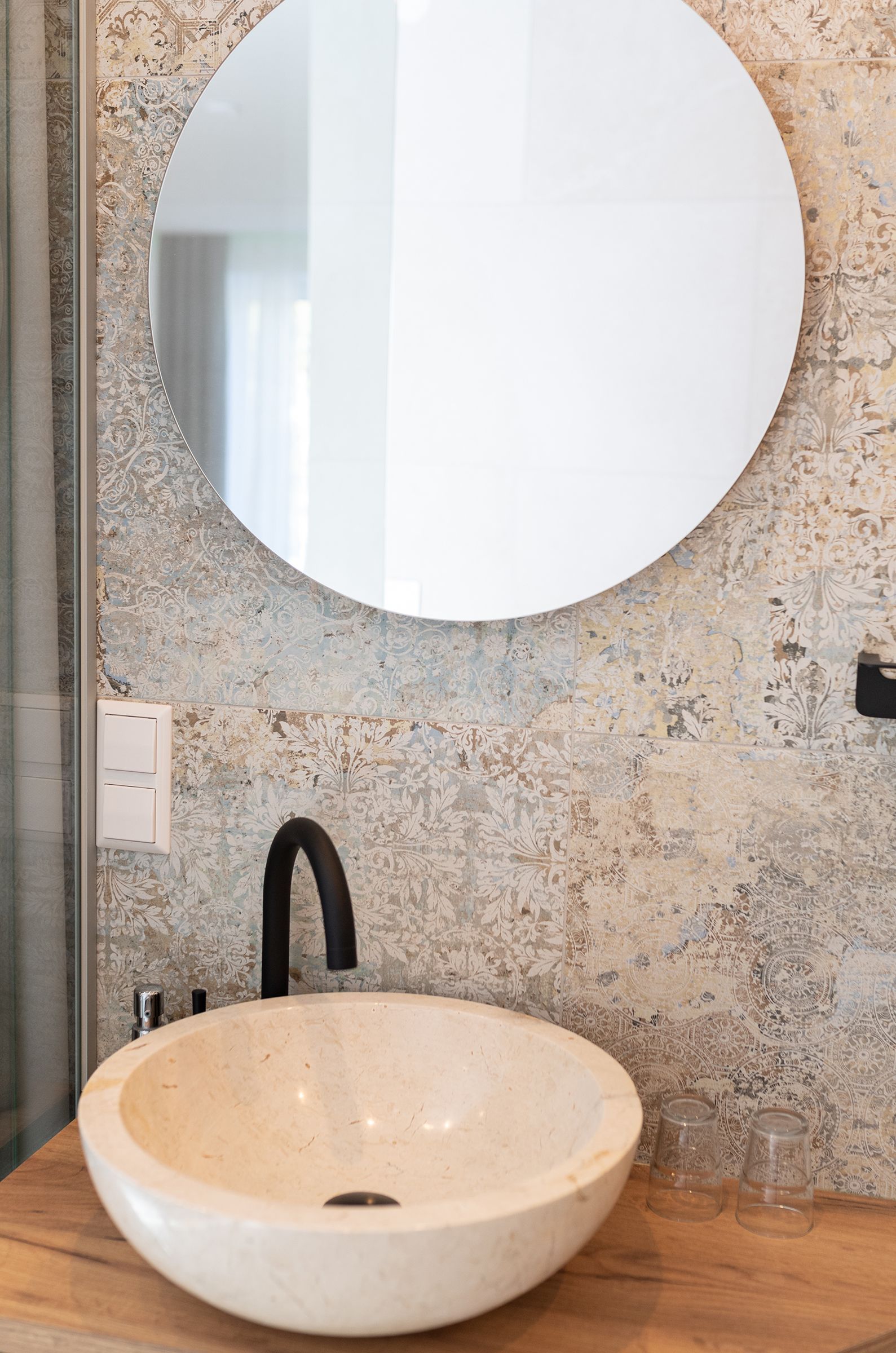 Ein rundes weißes Stein Waschbecken auf einem Holztisch mit schwarzer Armatur. darüber ist ein runder Spiegel. Die Rückwand ist mit strukturierten, beige-grauen Fliesen im Vintage-Dekor gestaltet