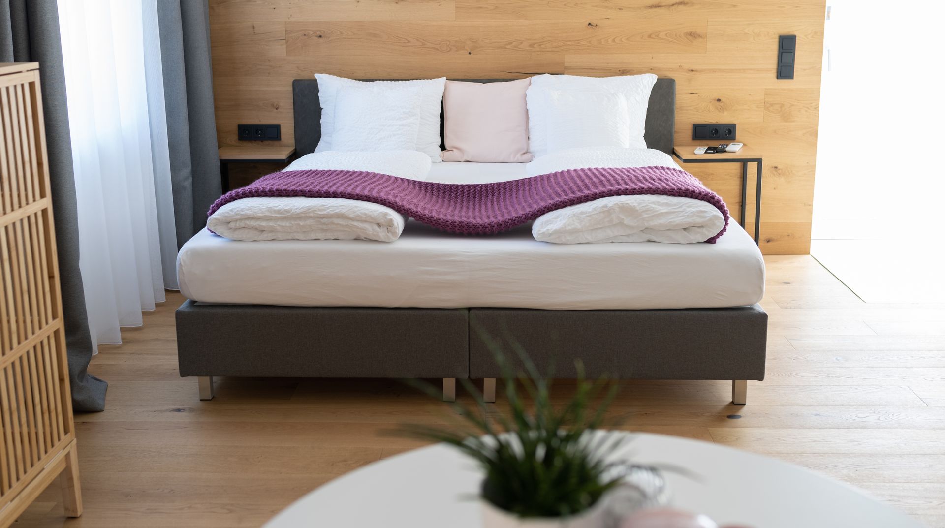 Ein graues Doppelbett mit weißen Laken steht im Fokus. Der Kopfteil des Bettes steht an einer Holzverkleideten Wand. Der Boden ist ebenso aus Parkett. Rechts und links neben dem Bett stehen Nachttische. Im Vordergrund ist ein weißer runder Tisch zu sehen mit einer Pflanze darauf.