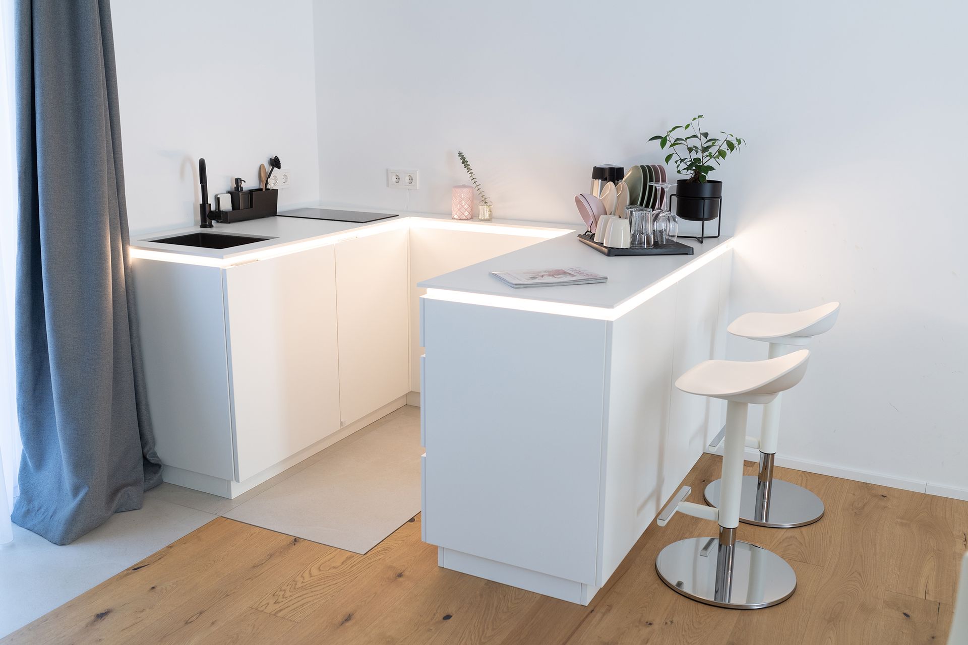 eine U-förmige weiße Küchenzeile mit indirekter Beleuchtung. darauf verschiedenes Geschirr und eine Pflanze. Ebenso ein Schwarzer Herd und Waschbecken. Die Küche ist neben dem großen Fenster.