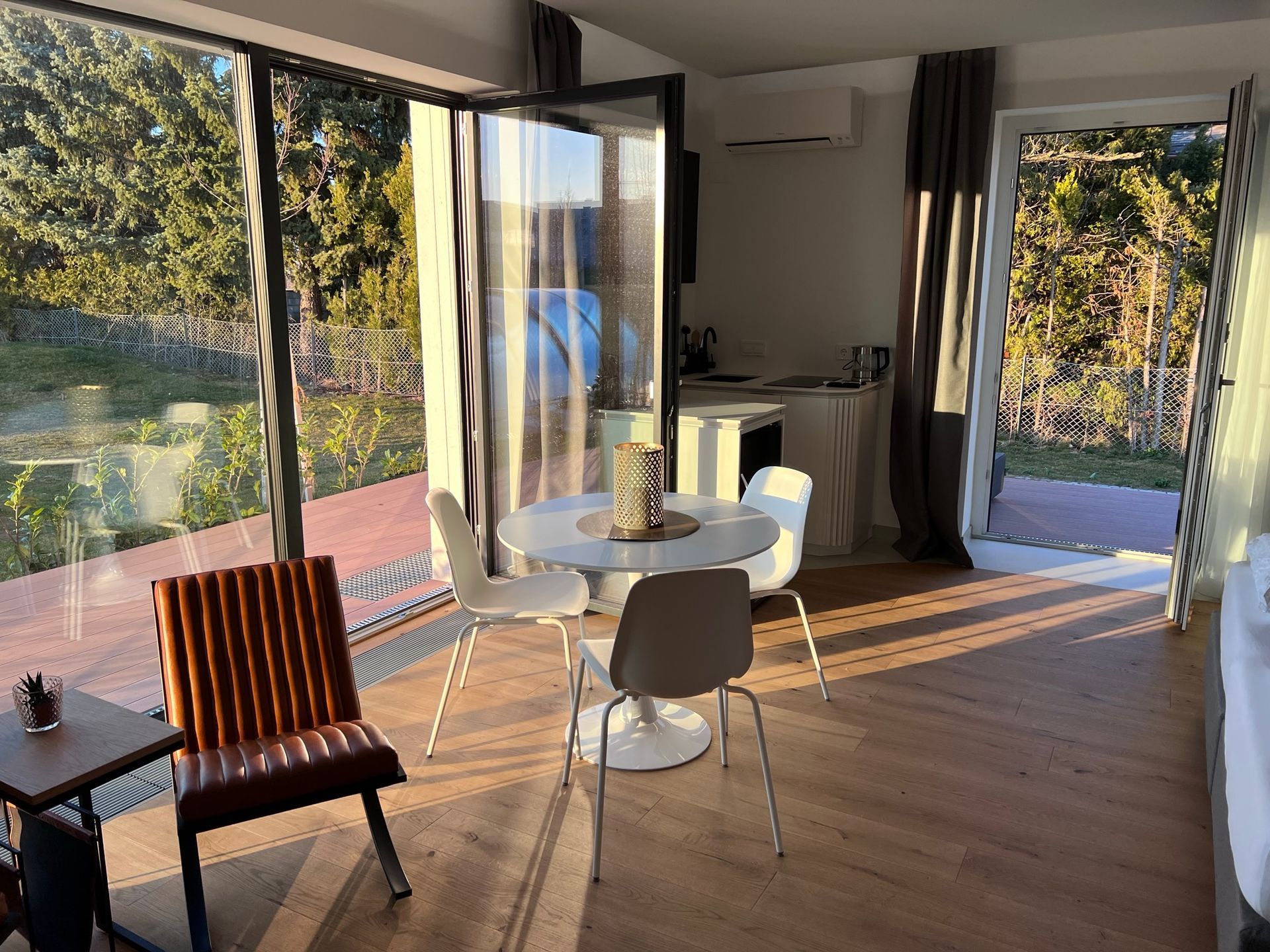 Zwei braune Lederstühle mit schwarzem Gestell aus Metall stehen an der Fensterwand. Dazwischen ein kleiner Beistelltisch. Dahinter der weiße, runde Esstisch mit passenden Stühlen. Daneben eine U-förmige weiße Küchenzeile. 