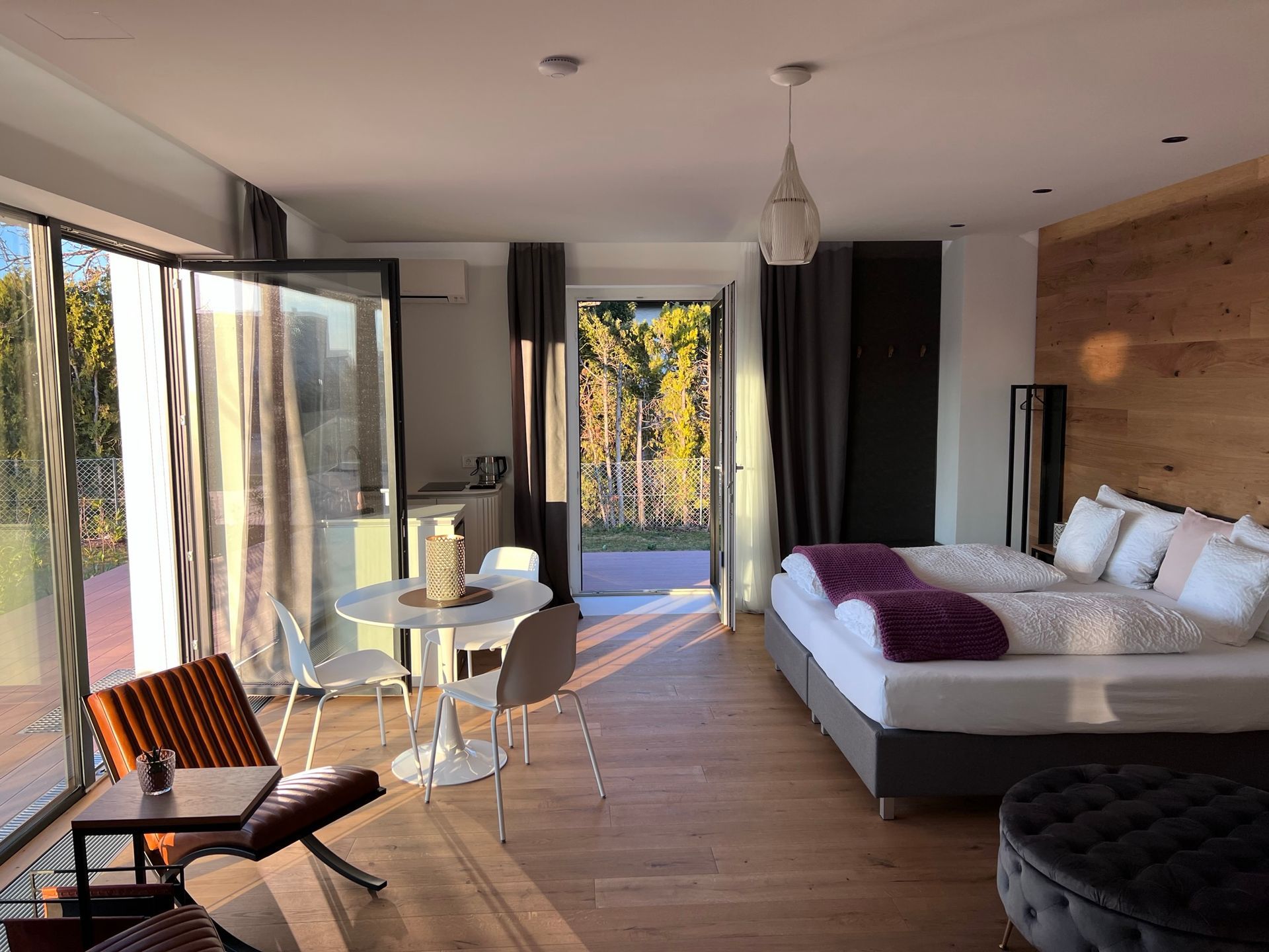 Modern eingerichtetes Zimmer mit großem Bett mit weißen Laken. Das Kopfteil steht an einer heller Holzwand. Gegenüber ist eine kleiner Sitzecke mit rundem Tisch und Stühlen sowie großen Fenstern. Durch die Fenster strahlt das Sonnenlicht hinein.