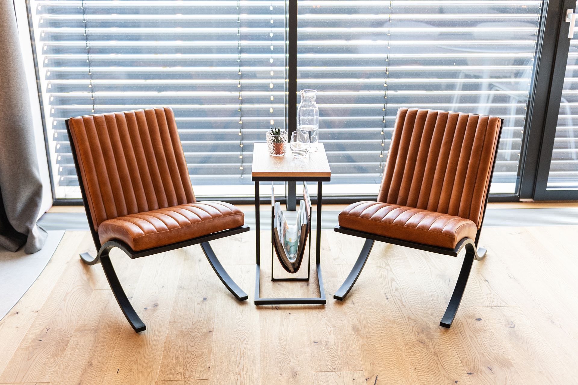 Zwei braune Lederstühle mit schwarzem Gestell aus Metall stehen an der Fensterwand. Dazwischen ein kleiner Beistelltisch. mit einer Wasserkaraffe darauf.