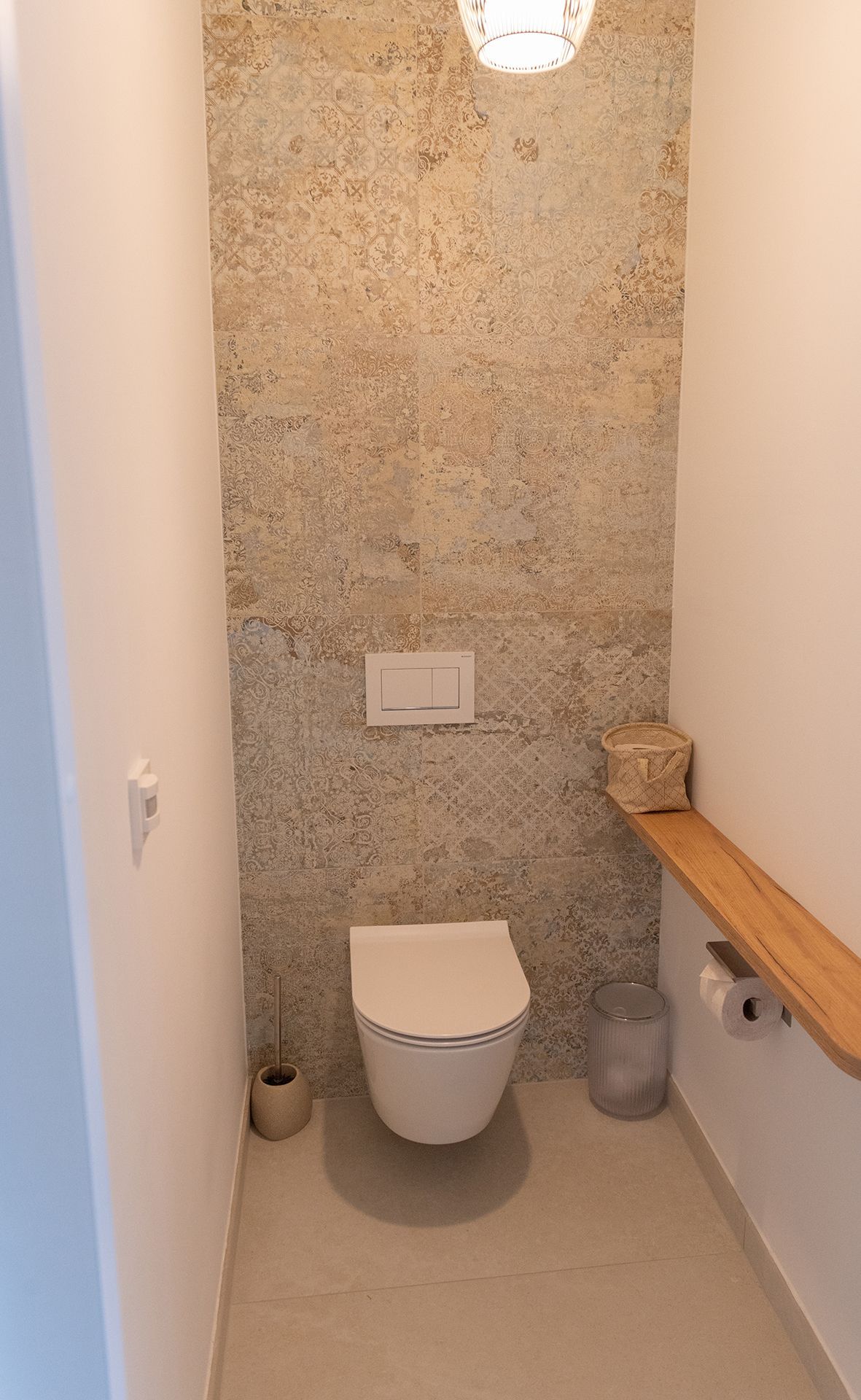 Modernes WC an einer verfliesten Wand. Daneben ist eine Ablage aus Holz. Neben dem WC ist ein kleiner Mistkübel und Klobesen.