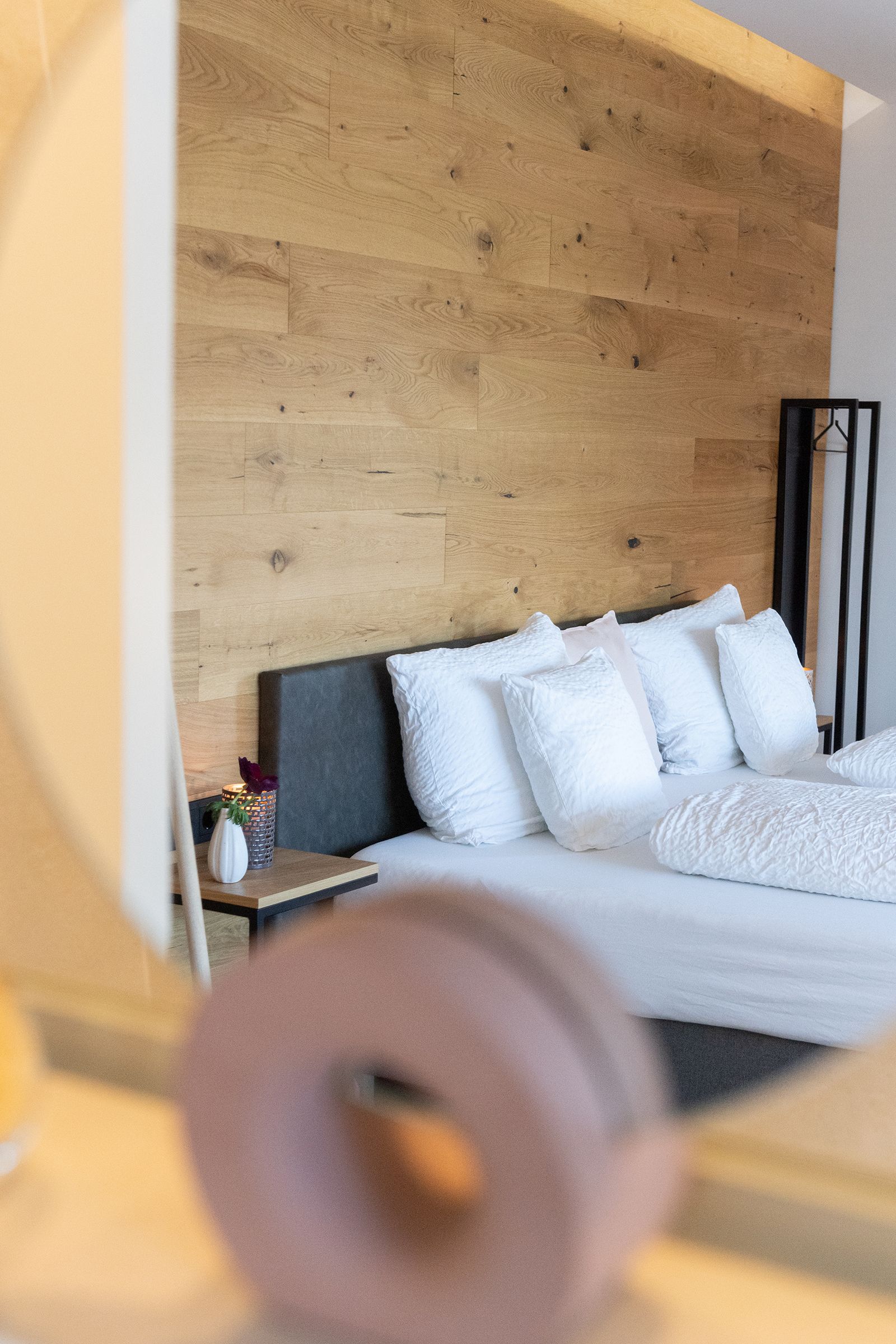 Das graue Doppelbett mit weißen Laken steht im Fokus. Links daneben ist ein kleiner Nachttisch mit Deko darauf. rechts daneben ist eine offene schwarze Garderobe aus Stahl.