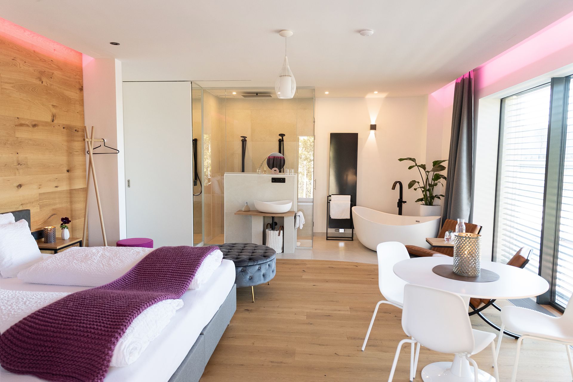 Modern eingerichtetes Zimmer mit offenem Badezimmerbereich, freistehender Badewanne, großem Bett mit weißen Laken. Das Kopfteil steht an einer heller Holzwand. Gegenüber ist eine kleiner Sitzecke mit rundem Tisch und Stühlen sowie großen Fenstern.