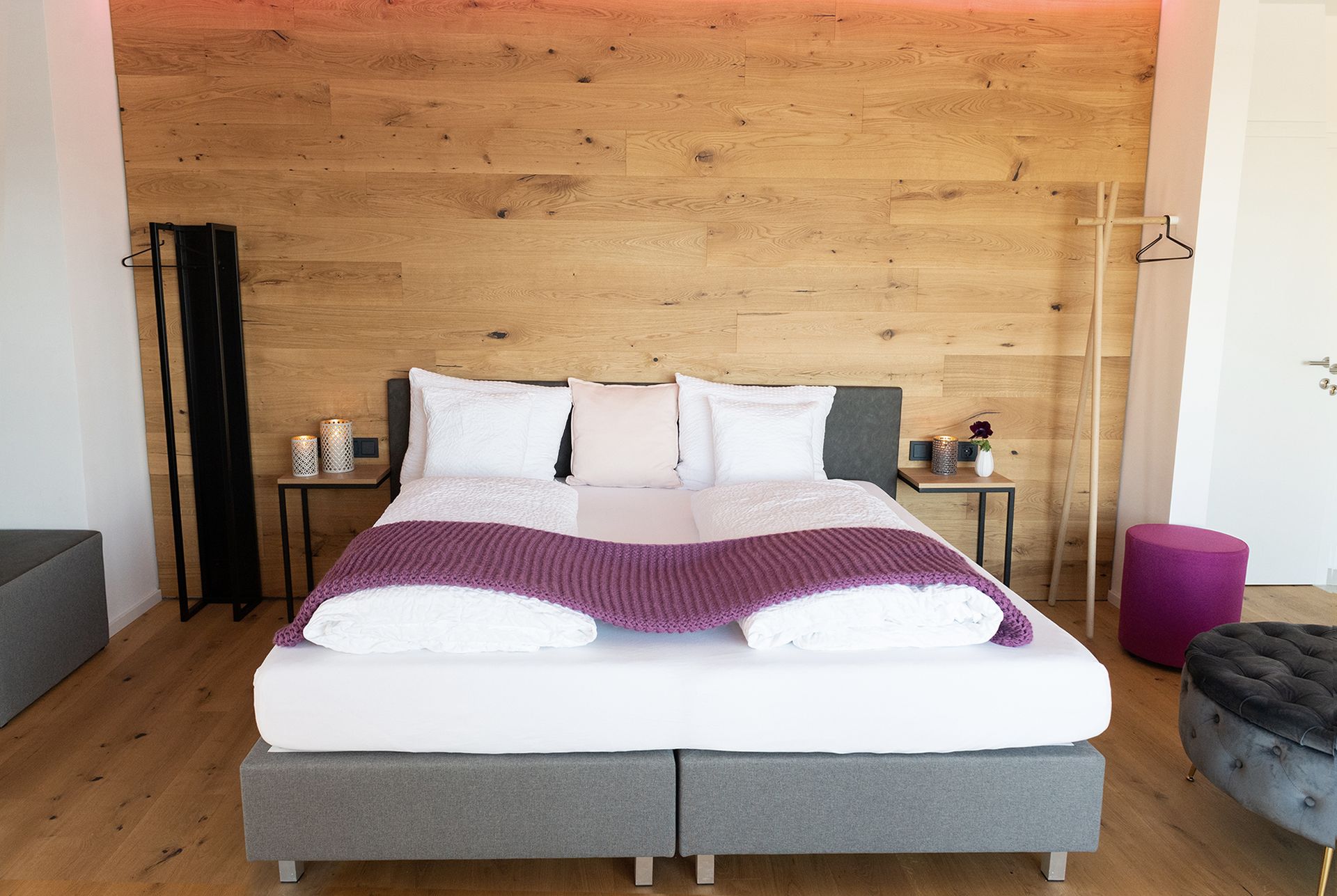 Das graue Doppelbett mit weißen Laken steht im Fokus. Links daneben ist ein kleiner Nachttisch mit Deko darauf. rechts daneben ist eine offene schwarze Garderobe aus Stahl.