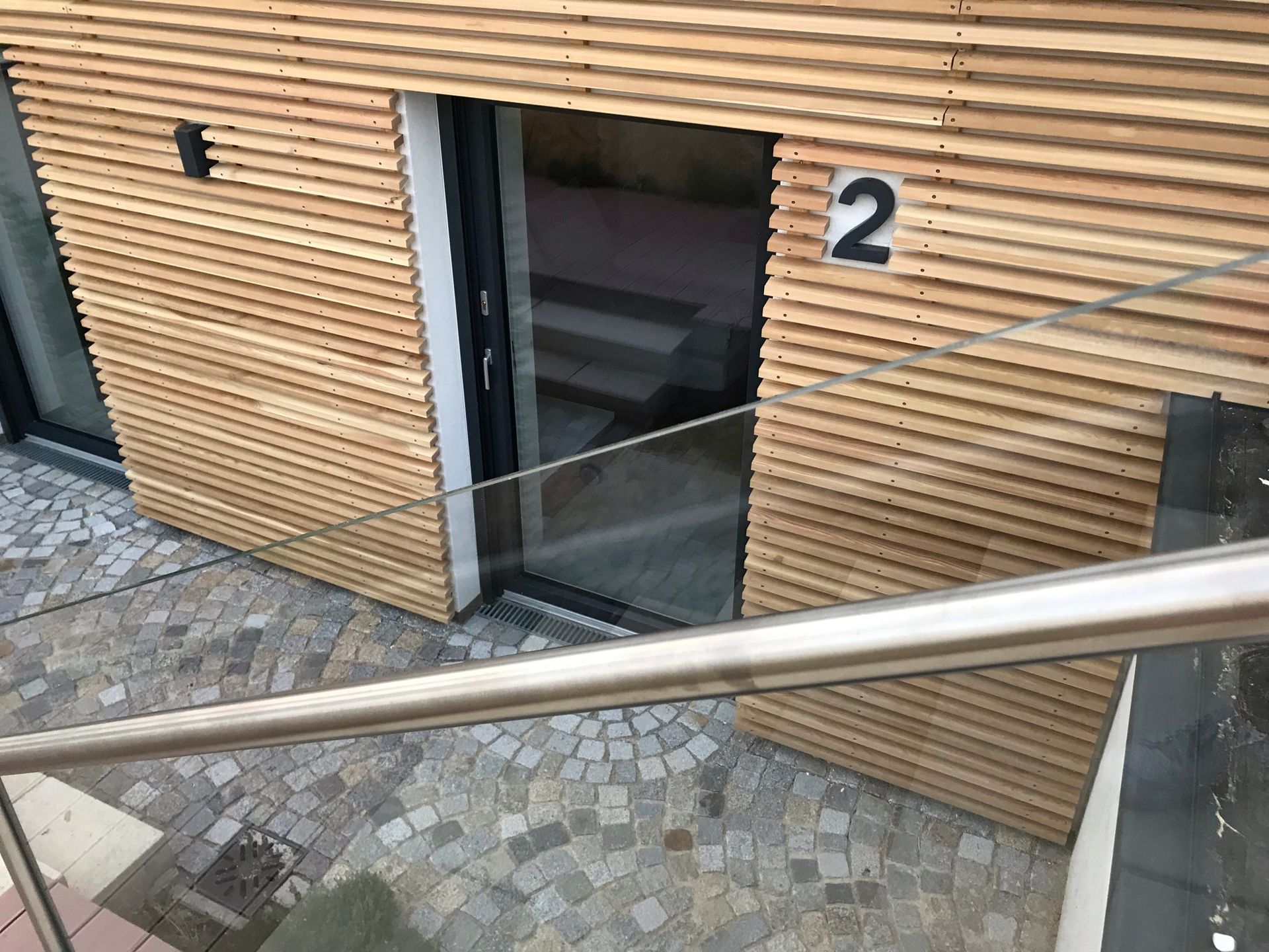 Ein Glasgelände führt nach unten. Der Eingang ist eingebettet in einer Wand, welche mit Holzpanelen verkleidet ist. Eine 