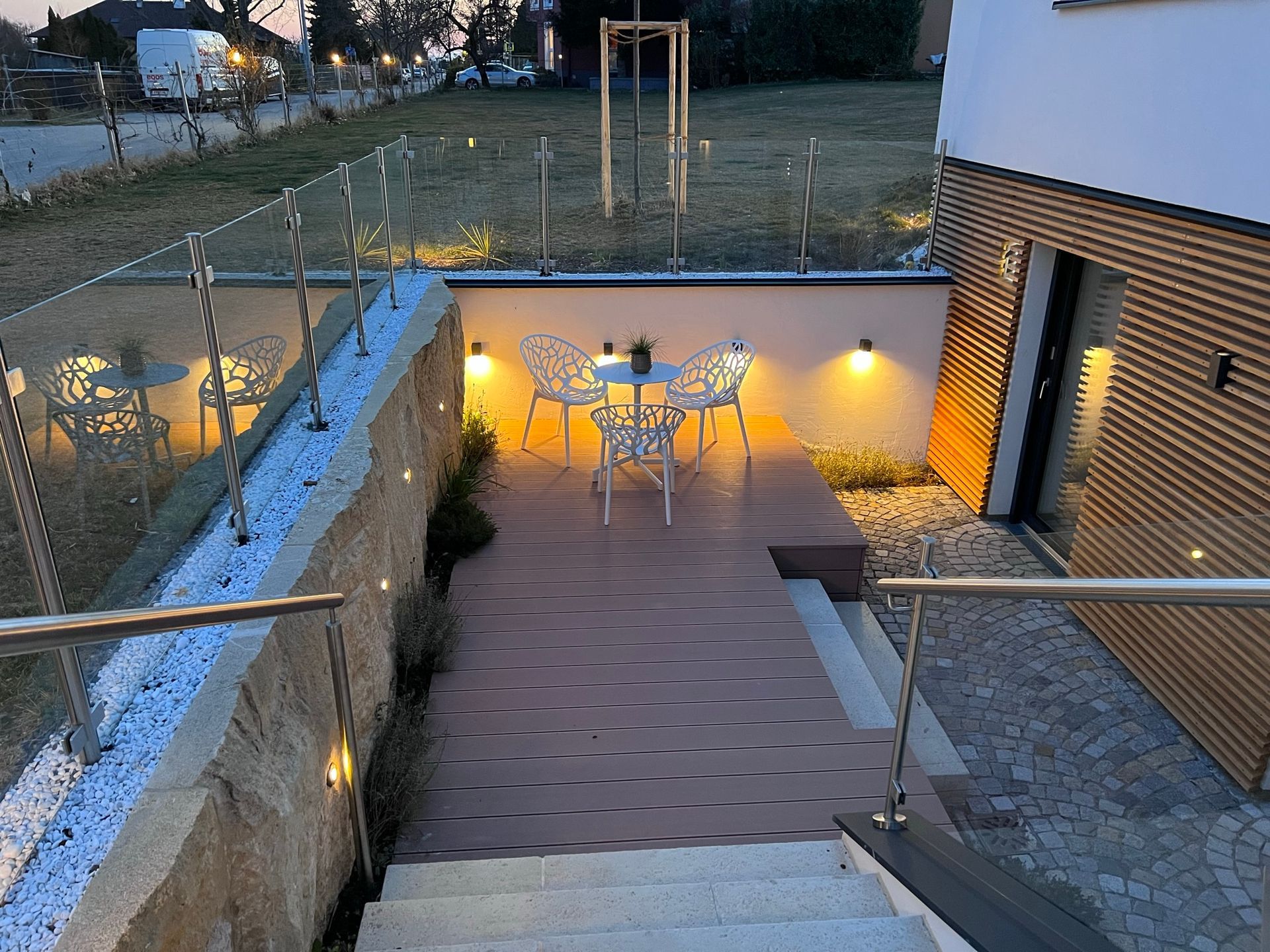 Stiegen führen hinunter aud die Terasse. Außenansicht der Terasse bei Dämmerung. Warme Beleuchtung an halbhoher Betonwand. Davor steht ein weißer, runder Tisch mit passenden Sesseln auf einer Holzterasse. Zwei Stiegen führen zum Eingang des Apartments.