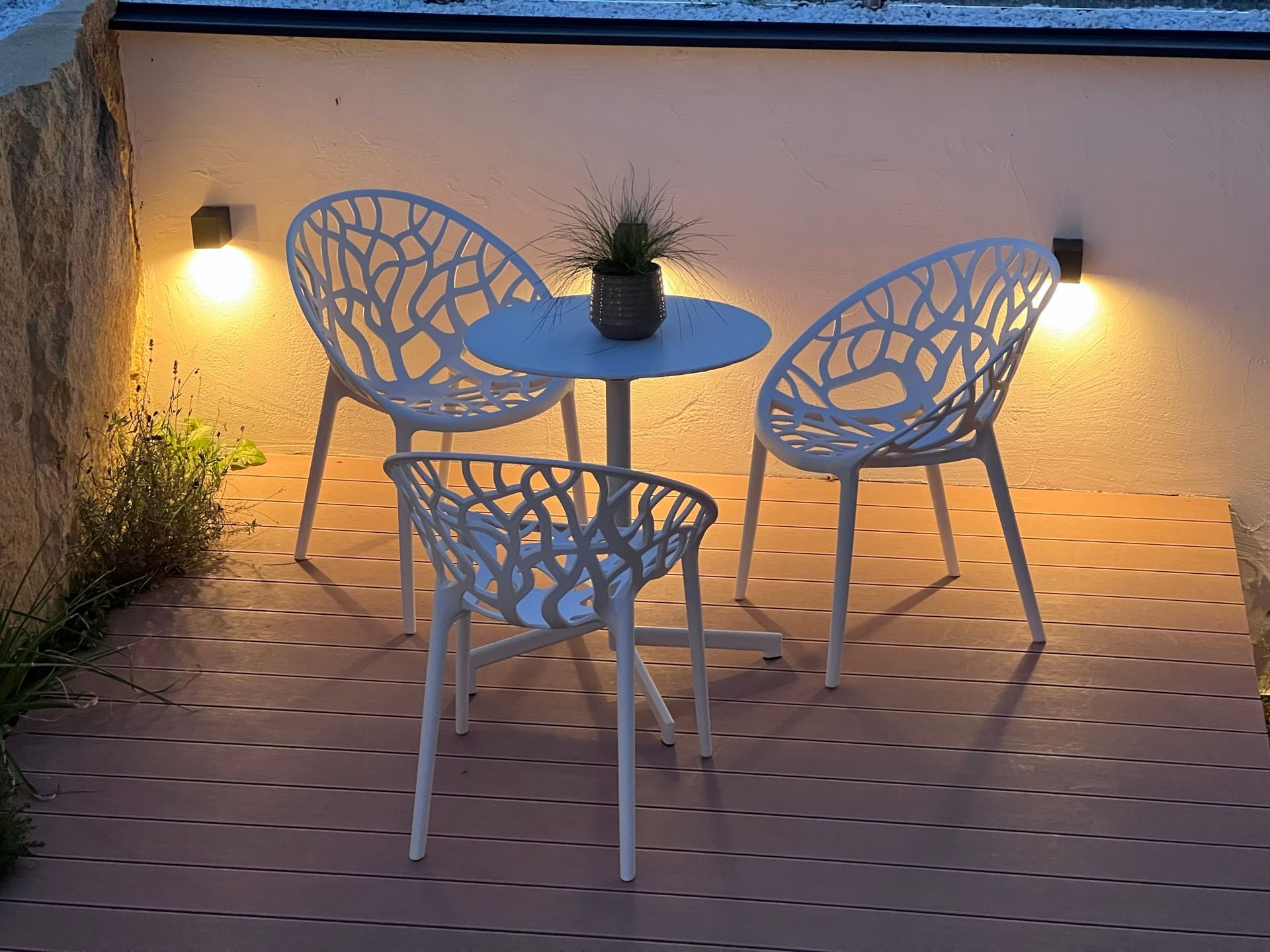 auf einer höhergelegten Terrasse mit Holzboden steht ein weißer runder Tisch und zwei weiße Stühle bei Dämmerung. Im Hintergrund ist eine beige Wand zu sehen. Die Wand ist beleuchtet.