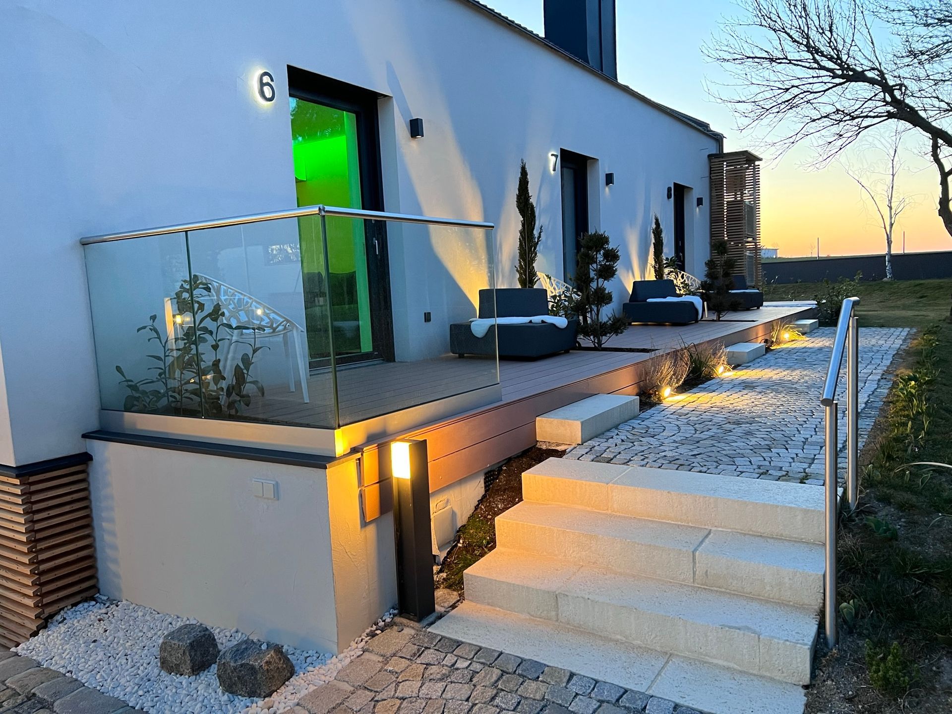 Außenansicht der Holzterrassen von Apartment 6, 7 und 8 bei Dämmerung. Jede Terrasse hat einen grauen Stoffsessel und Pflanzen. Die weiße Wand und die grauen Fenstertüren bilden einen Kontrast. Der Weg und die Terrassen sind beleuchtet.