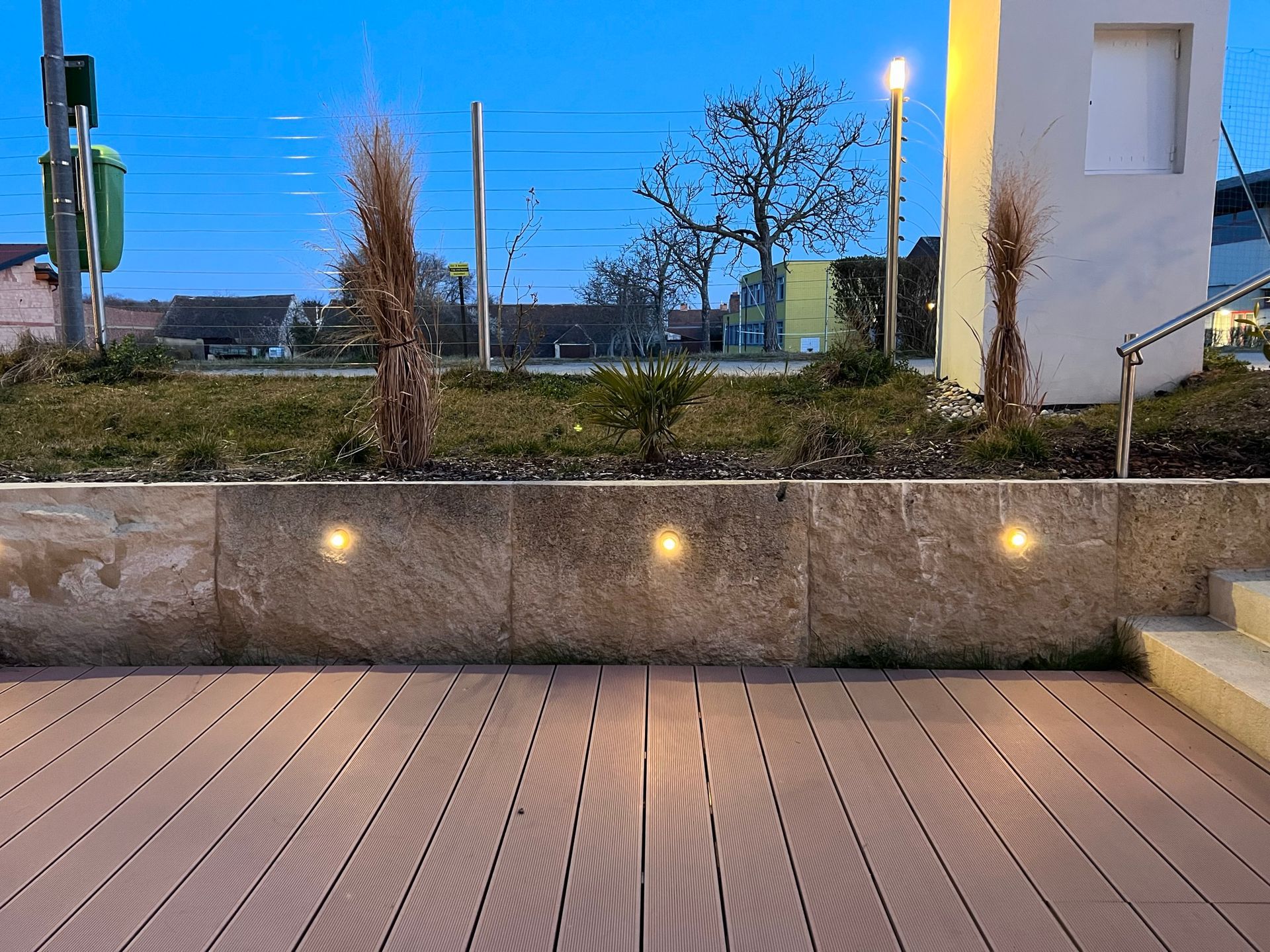 auf einer höhergelegten Terrasse mit Holzboden steht ein weißer runder Tisch und zwei weiße Stühle bei Dämmerung. Im Hintergrund ist eine halbhohe Steinwand und der Himmel zu sehen. Die Wand ist beleuchtet.