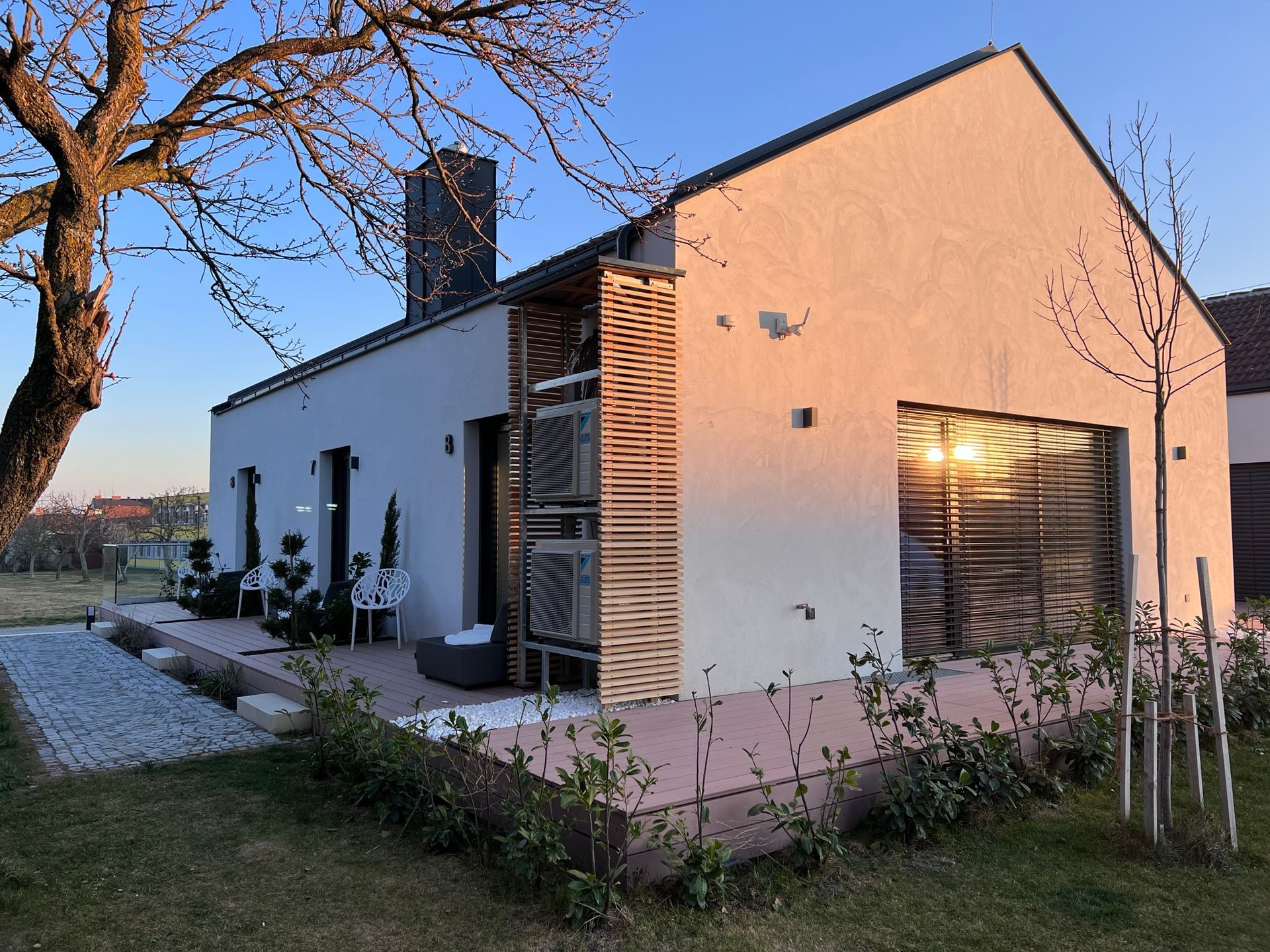 Außenansicht der Holzterrassen von Apartment 6, 7 und 8. Jede Terrasse hat einen grauen Stoffsessel und Pflanzen. Der Sonnenuntergang spiegelt sich im Fenster von Apartment 8. Ein Baum ragt von der Seite in das Bild hinein.