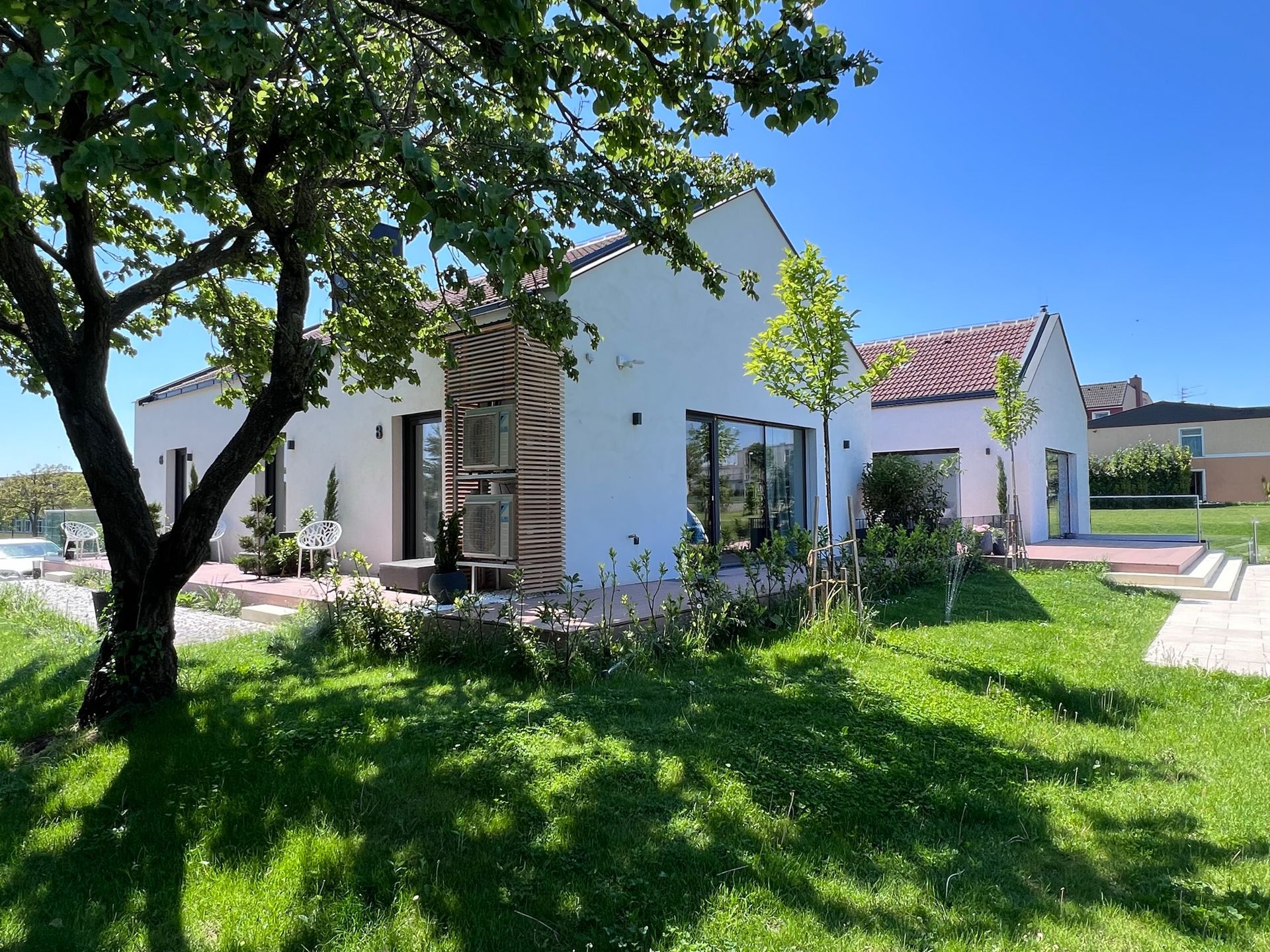 Außenansicht der Holzterrassen von Apartment 6, 7 und 8. Jede Terrasse hat einen grauen Stoffsessel und Pflanzen. Der Rasen ist gepflegt und grün. Ein Baum ragt von der Seite in das Bild hinein.
