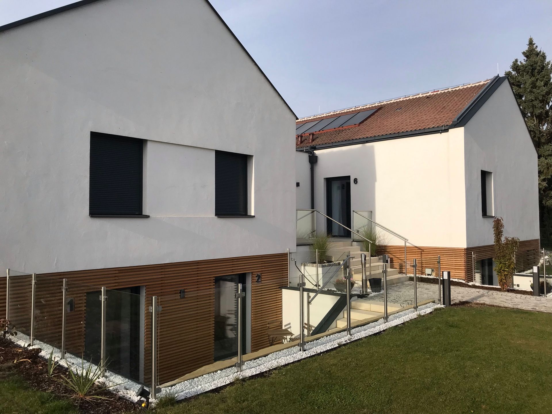 Außenansicht frontal bei Sonnenaufgang. Großes weißes haus mit Holzelementen. ein Pflasterstein Weg und Stiegen hinunter führen zu Appartment 1, 2.
Der angrenzende Rasen ist gepflegt.
