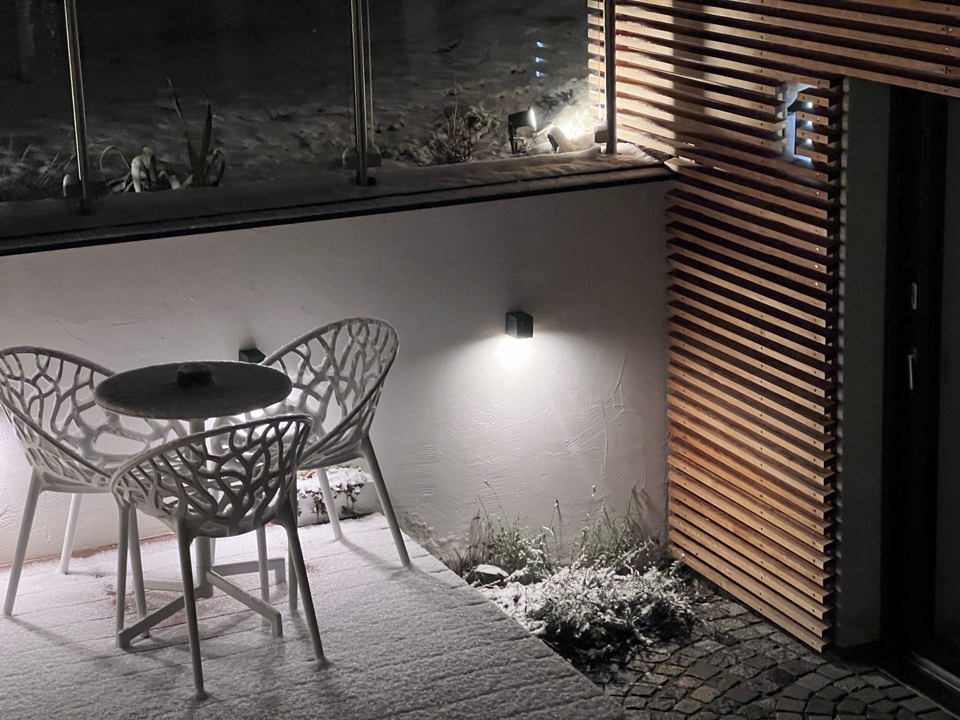 auf einer höhergelegten Terrasse mit Holzboden steht ein weißer runder Tisch und zwei weiße Stühle bei Dämmerung. Im Hintergrund ist eine beige Wand und teil der Holzfassade zu sehen. Die Wand ist beleuchtet. Auf Boden, Sessel und Tisch ist Schnee.