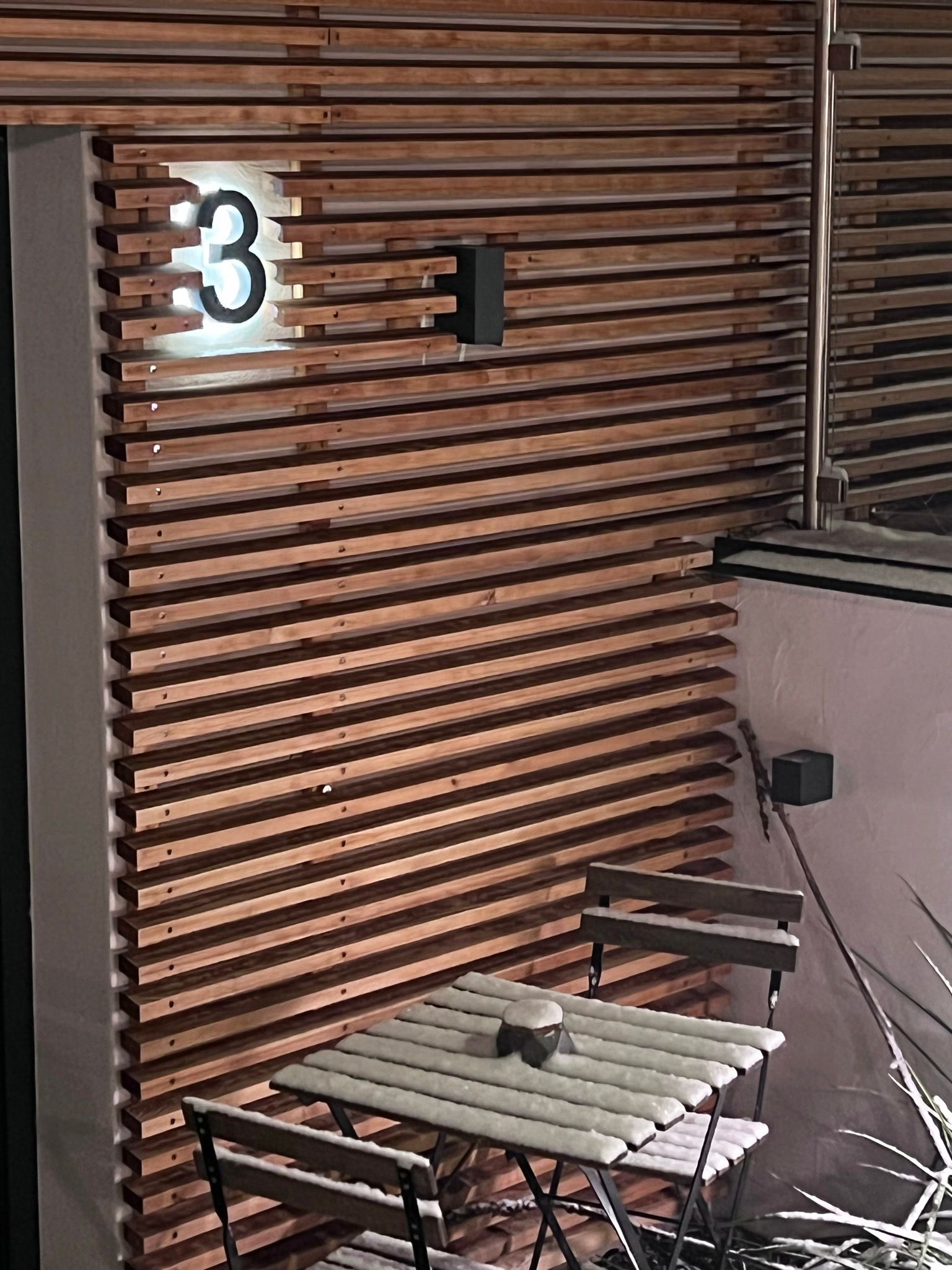 Außenbereich mit Holzterrasse, einem Holztisch sowie zwei passende Stühle. Indirekte Beleuchtung findet sich in der Holzfassade an der Wand des Eingangs zu Apartment 3. Der kleine Tisch ist mit Schnee bedeckt.