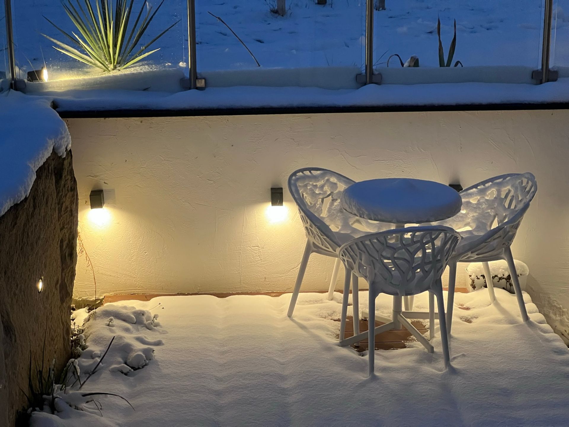 auf einer höhergelegten Terrasse mit Holzboden steht ein weißer runder Tisch und zwei weiße Stühle bei Dämmerung. Im Hintergrund ist eine beige Wand zu sehen. Die Wand ist beleuchtet. Auf Boden, Sessel und Tisch ist Schnee.