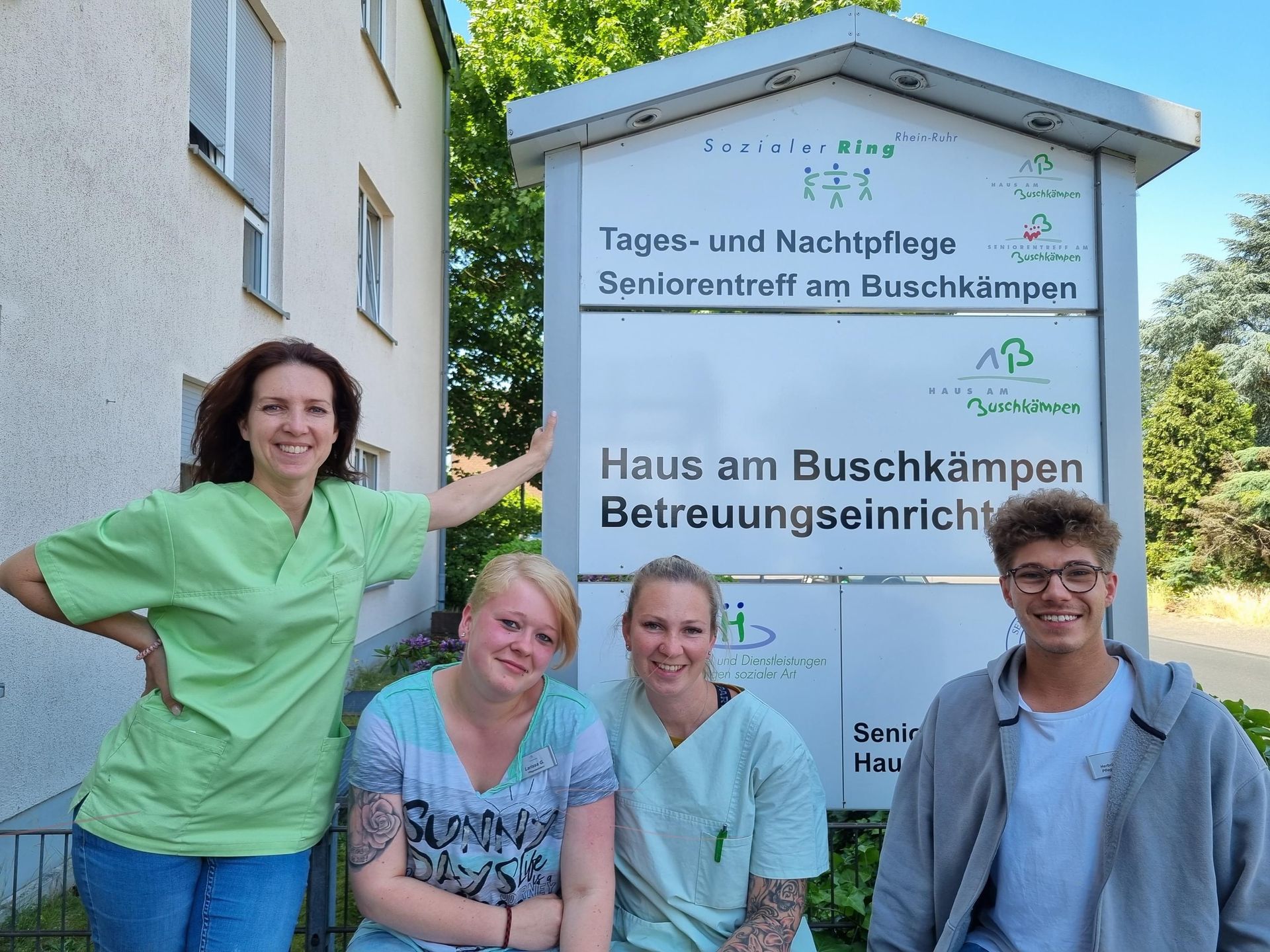 Mehrere Mitarbeitende blicken in Kamera, sie stehen am Eingangsschild zum Haus am Buschkämpen
