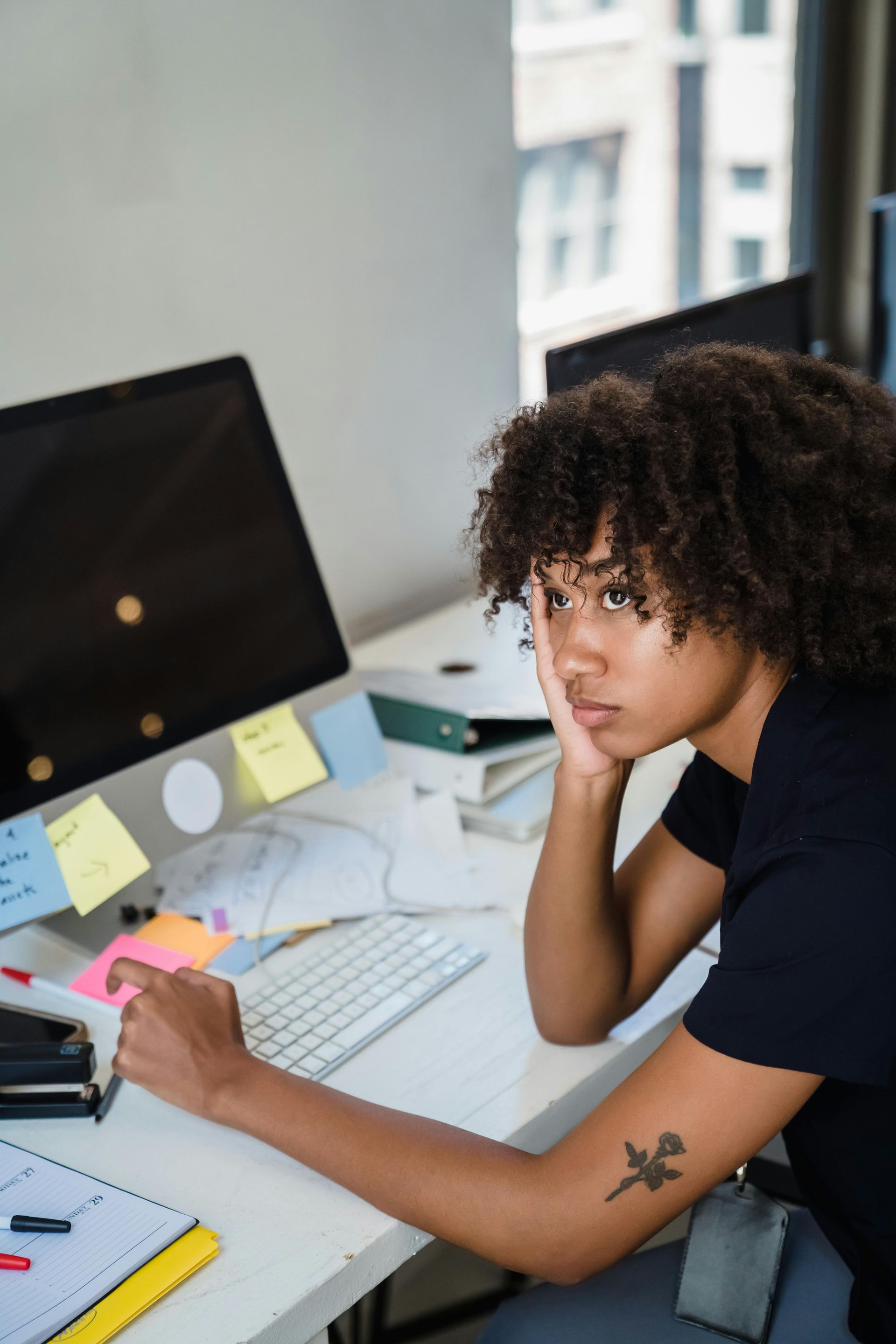 Woman at a desk, looking bored. A computer and paperwork are present. She has a tattoo.