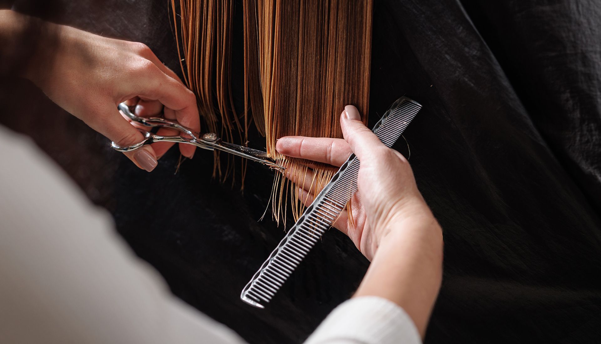 Hands using scissors and a comb to cut wet, reddish-brown hair in a salon setting.