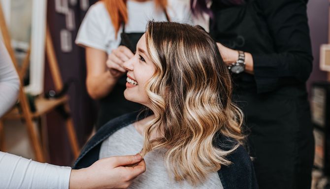 Woman smiles in a salon chair as stylists work on her wavy, highlighted hair.