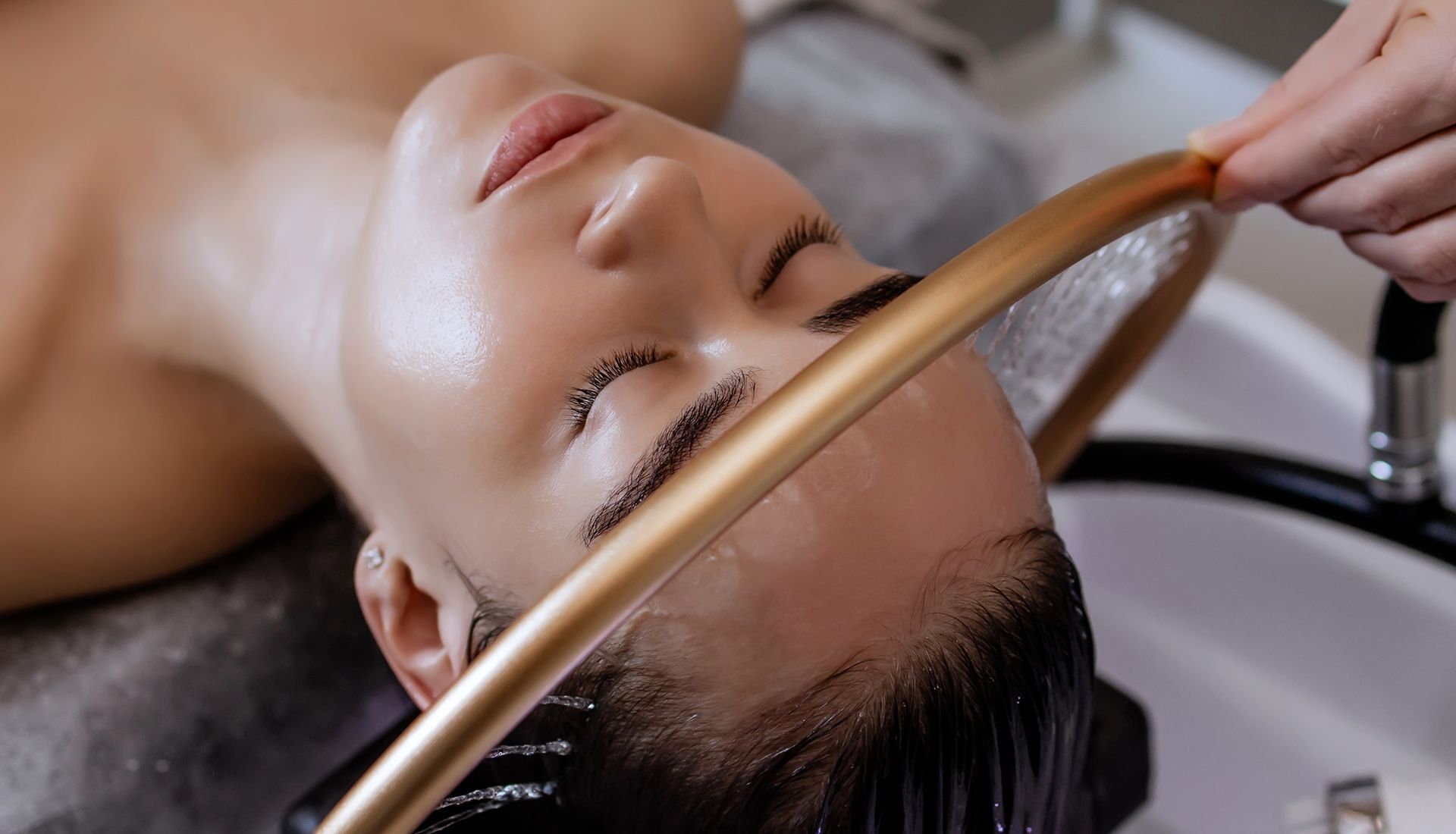 Person with closed eyes has hair rinsed at a salon sink; tan hose sprays water.