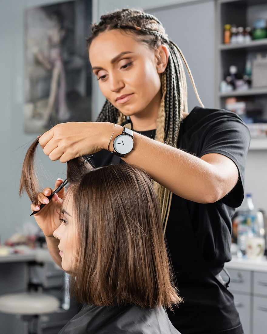 Hairdresser cutting a client's hair in a salon; client is seated. The hairdresser has braided hair, wearing a watch.