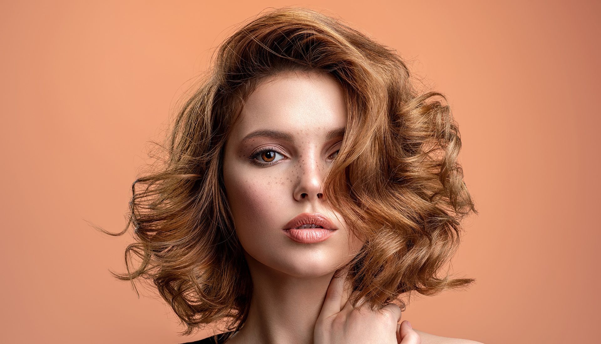 Woman with wavy, auburn hair, looking at the camera. Light skin, freckles, soft makeup, peach backdrop.