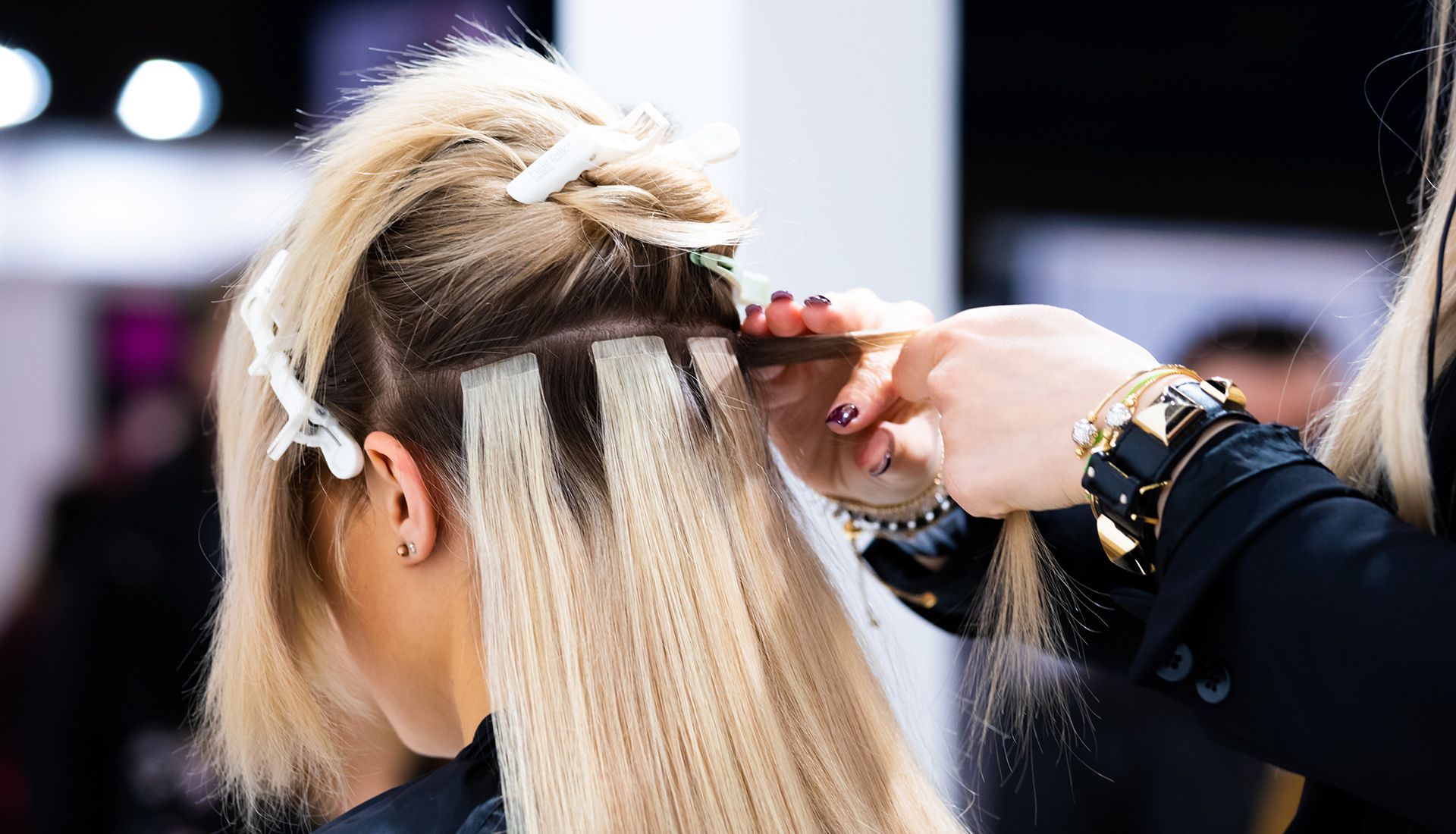 A stylist attaching blonde hair extensions to a client's natural hair with clips and tools.