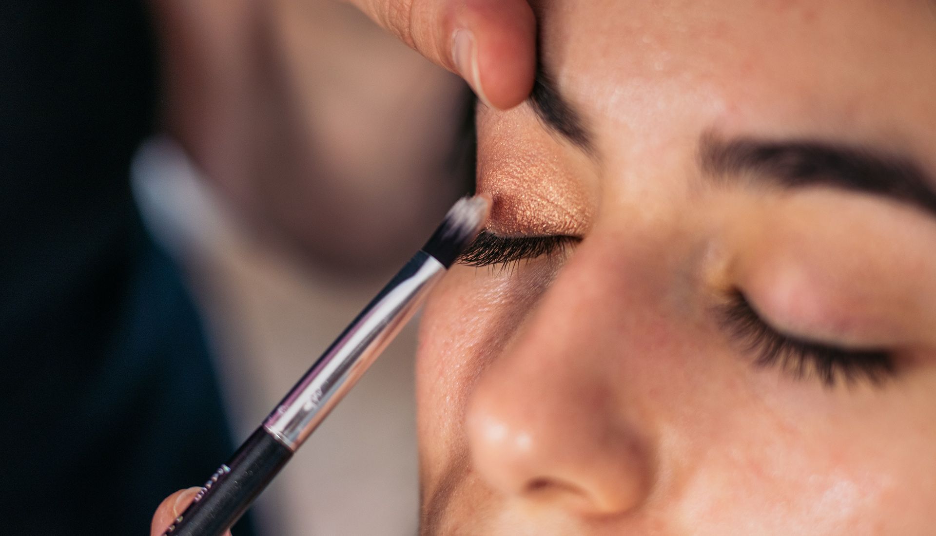 Makeup artist applying eyeshadow to a person's closed eyelid with a brush.