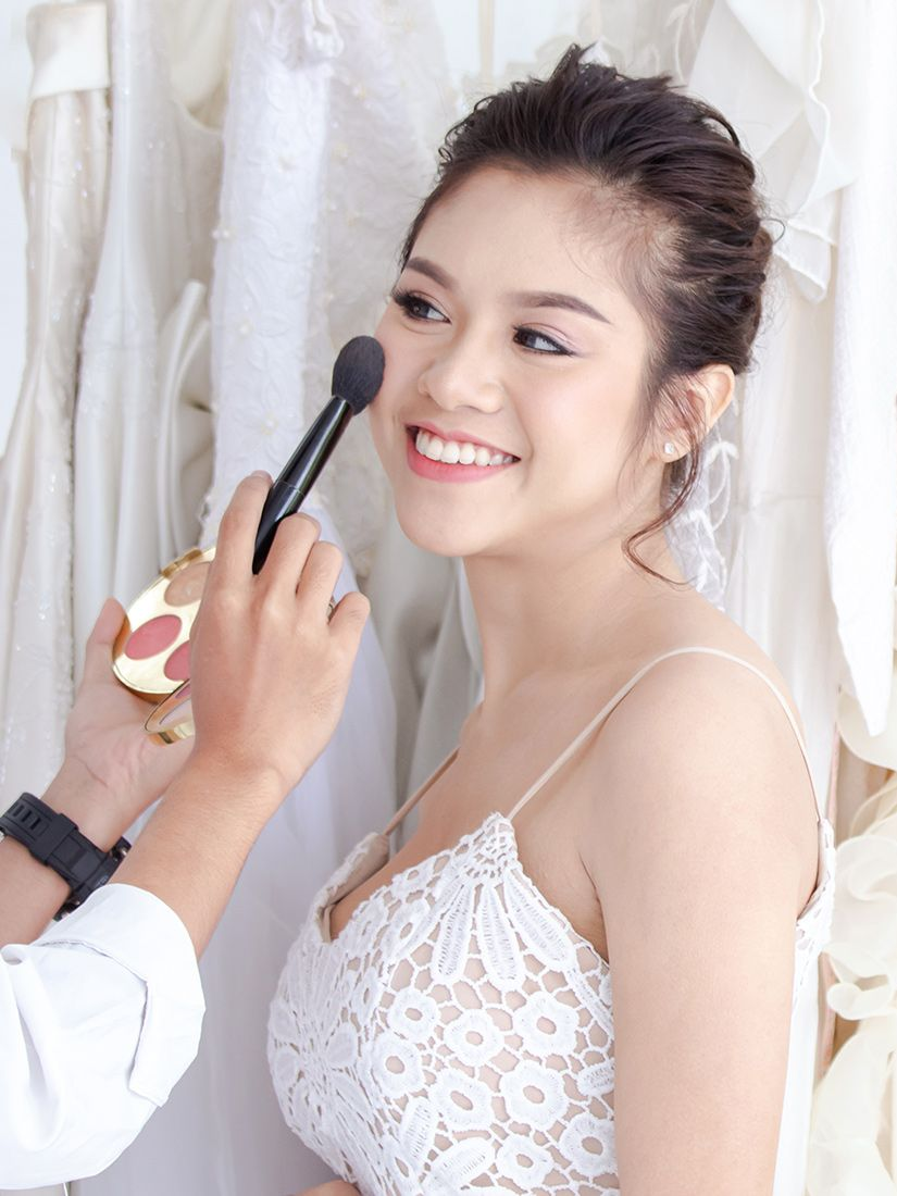 Makeup artist applying blush to a smiling person in a white lace dress. Wedding dress backdrop.