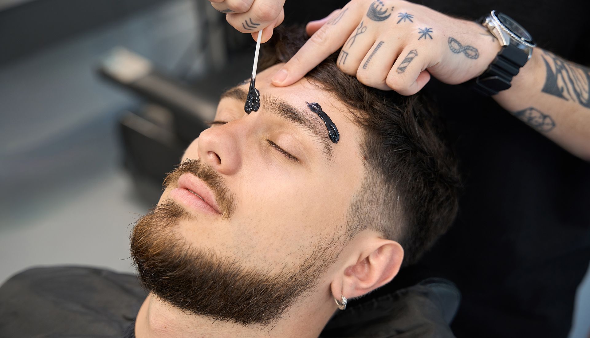 Man having eyebrow wax treatment. Technician applying wax with a stick. Man's eyes are closed, in a salon setting.