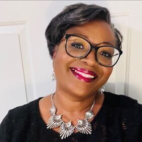 Woman with glasses, short gray hair, and necklace smiles, indoors.
