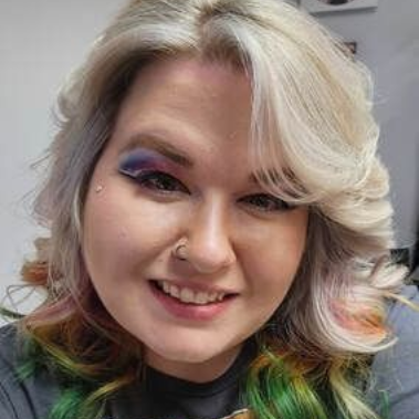 Woman with colorful hair and eye makeup, smiling, wearing a nose ring.