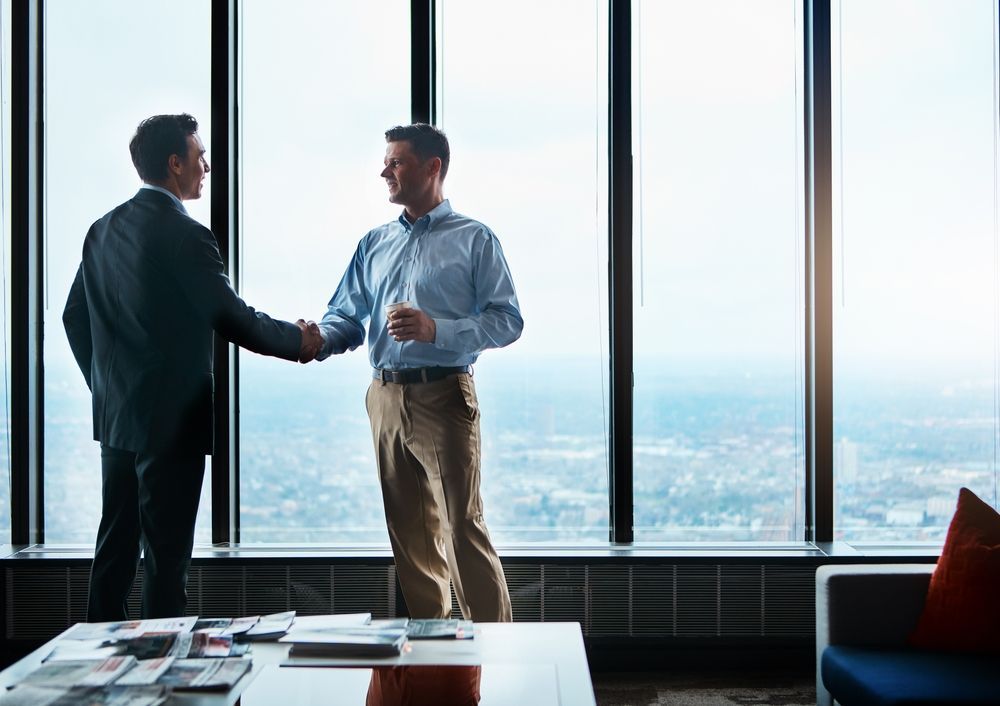 Two men are shaking hands in front of a large window.